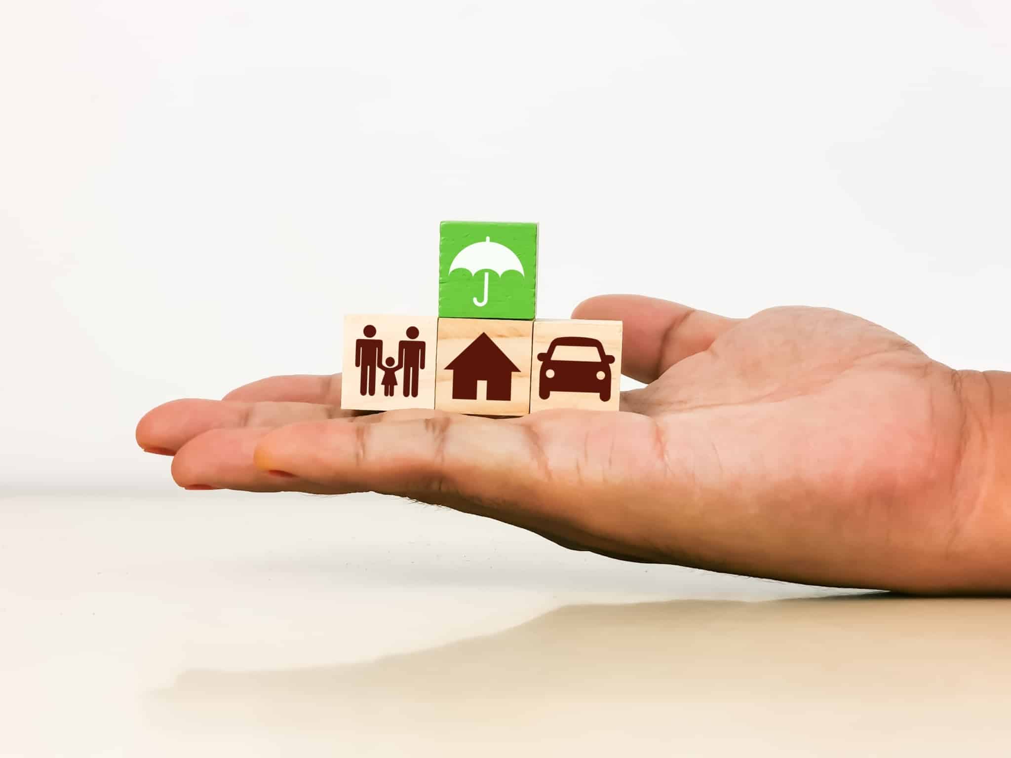 Mão a segurar peças de madeira com símbolos família, casa, automóvel e chapéu de chuva para simbolizar seguro de responsabilidade civil