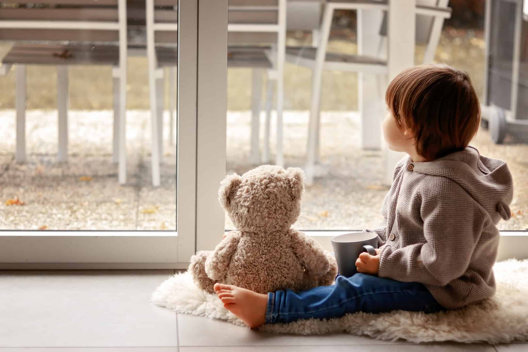 Criança sentada no chão com um urso de peluche ao lado, em frente a uma janela