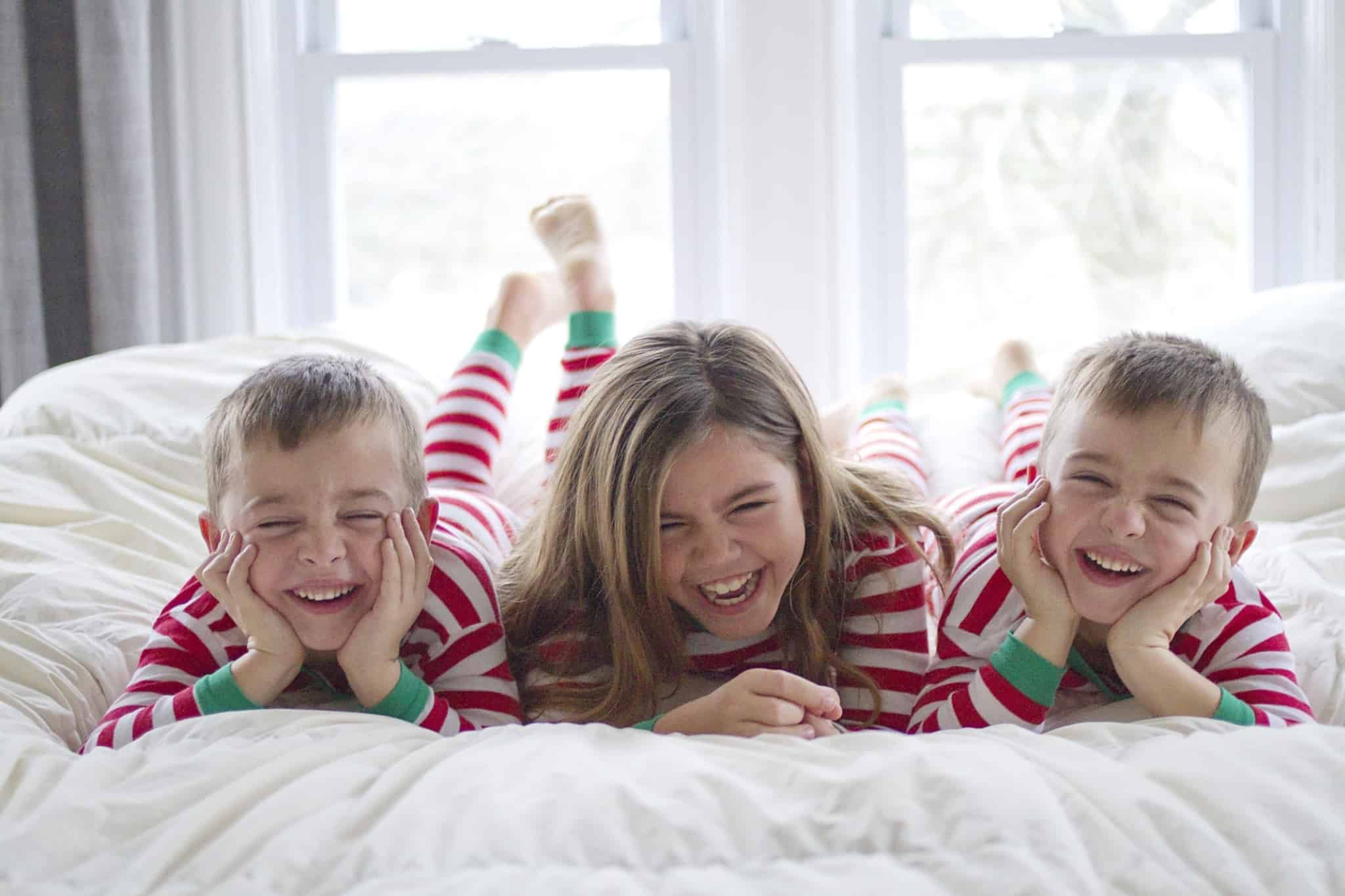 Três crianças deitadas na cama dos pais, de pijamas de Natal, com rasgados sorrisos