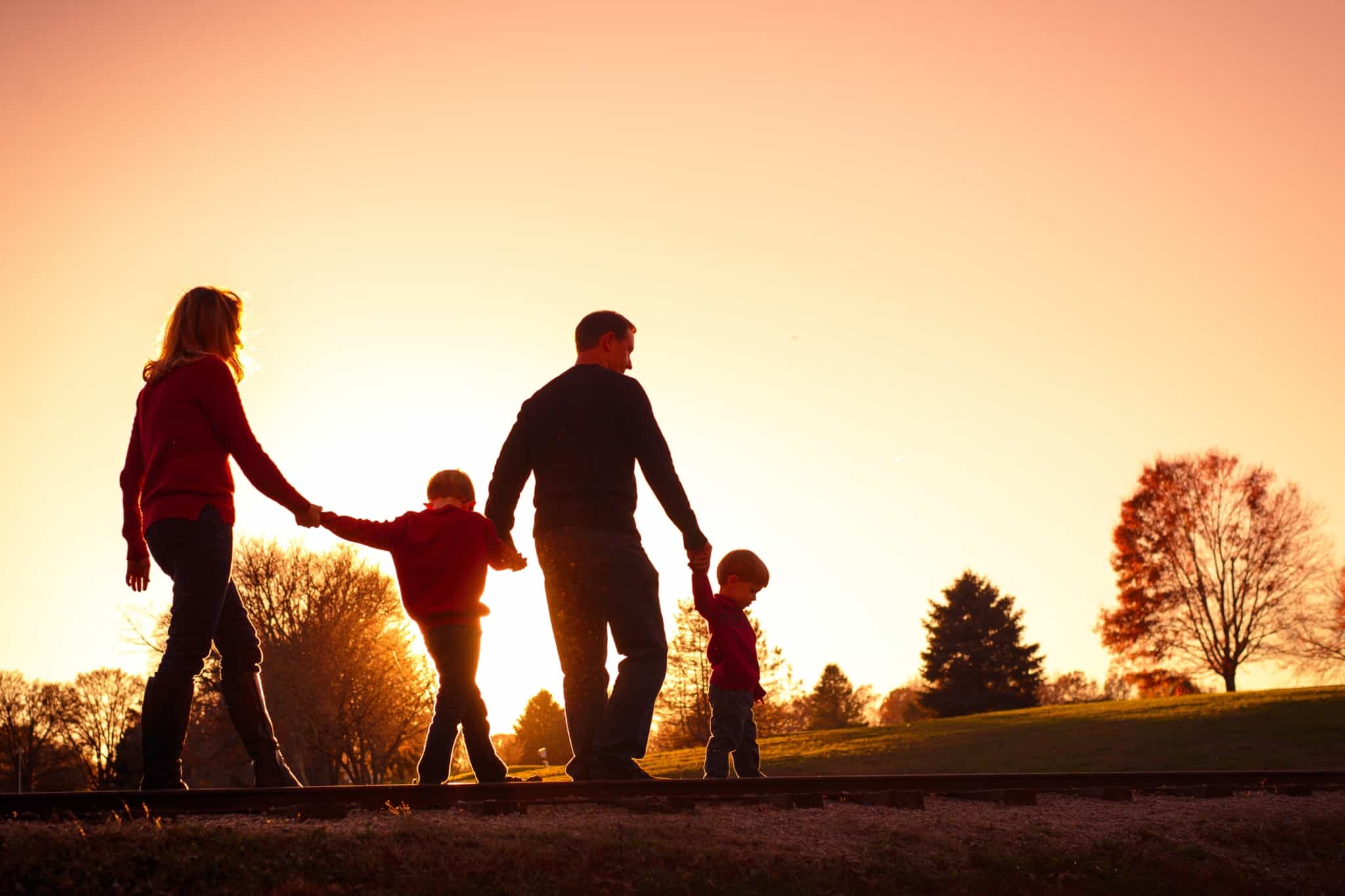 pai e mãe caminham de mãos dadas, entre a natureza, com os dois filhos pequenos com o pôr de sol no horizonte