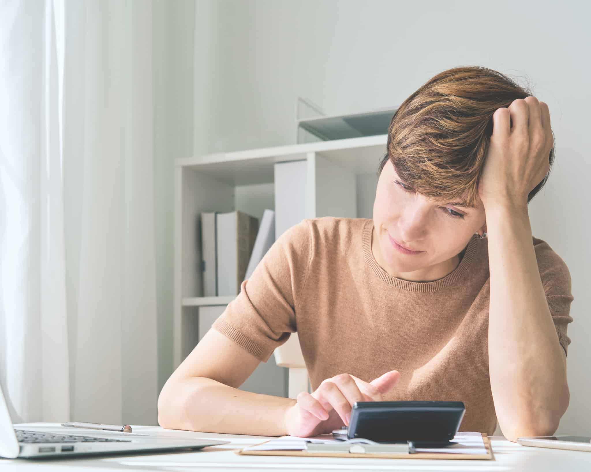 jovem mulher com a mão na cabeça e olhar preocupado faz contas, às suas dívidas, na calculadora