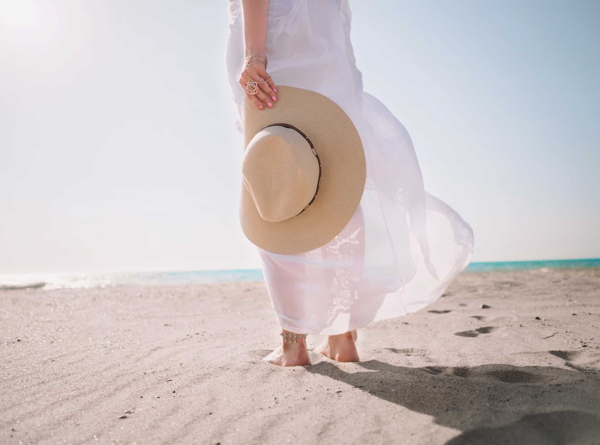 mulher caminha na areia da praia, num dia de muito sol, com um chapeu de palaha na mão