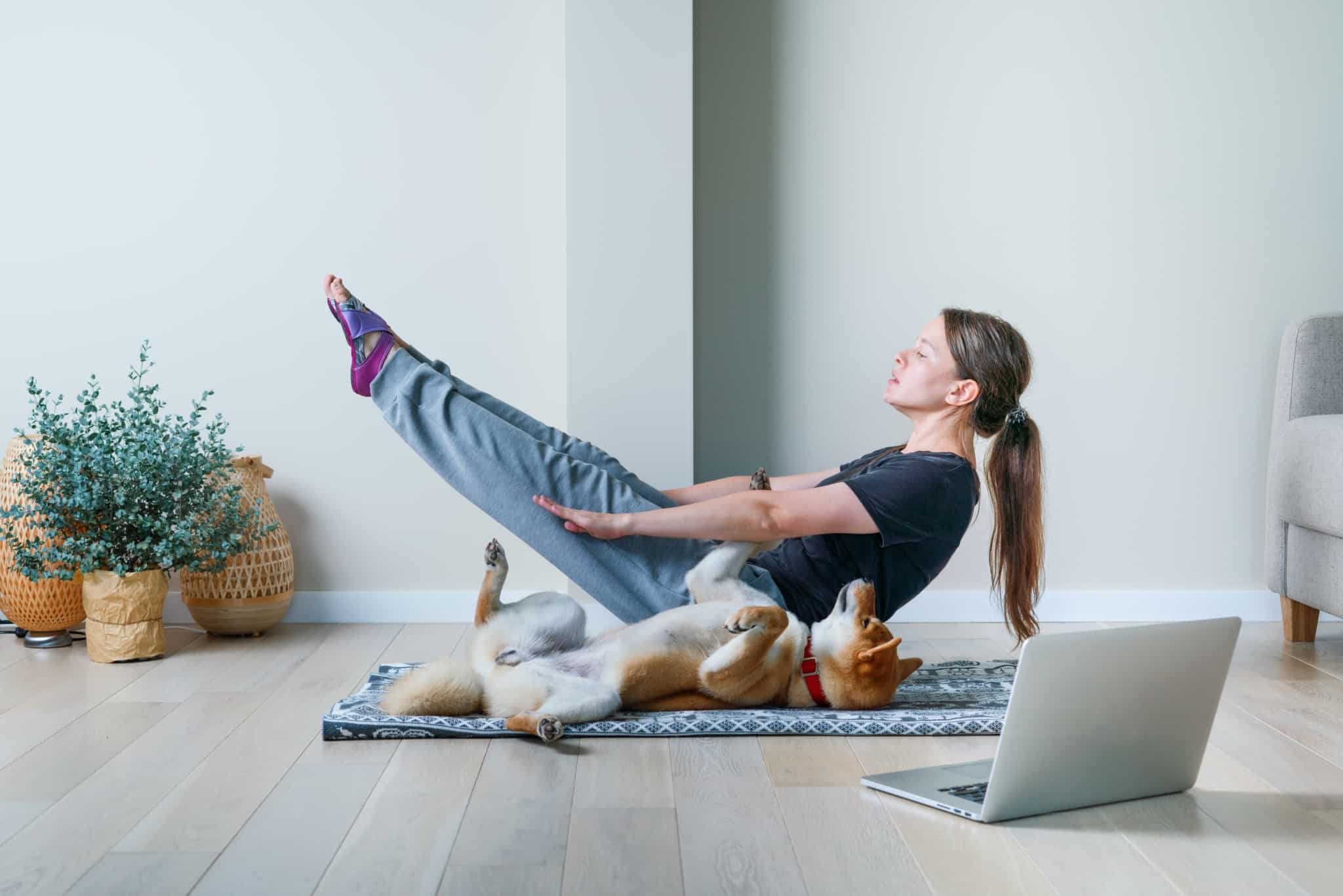 jovem mulher a fazer ioga, em casa, em posição de equil~ibroio de pernas erguidas e o seu cão ao lado a reproduzir o movimento