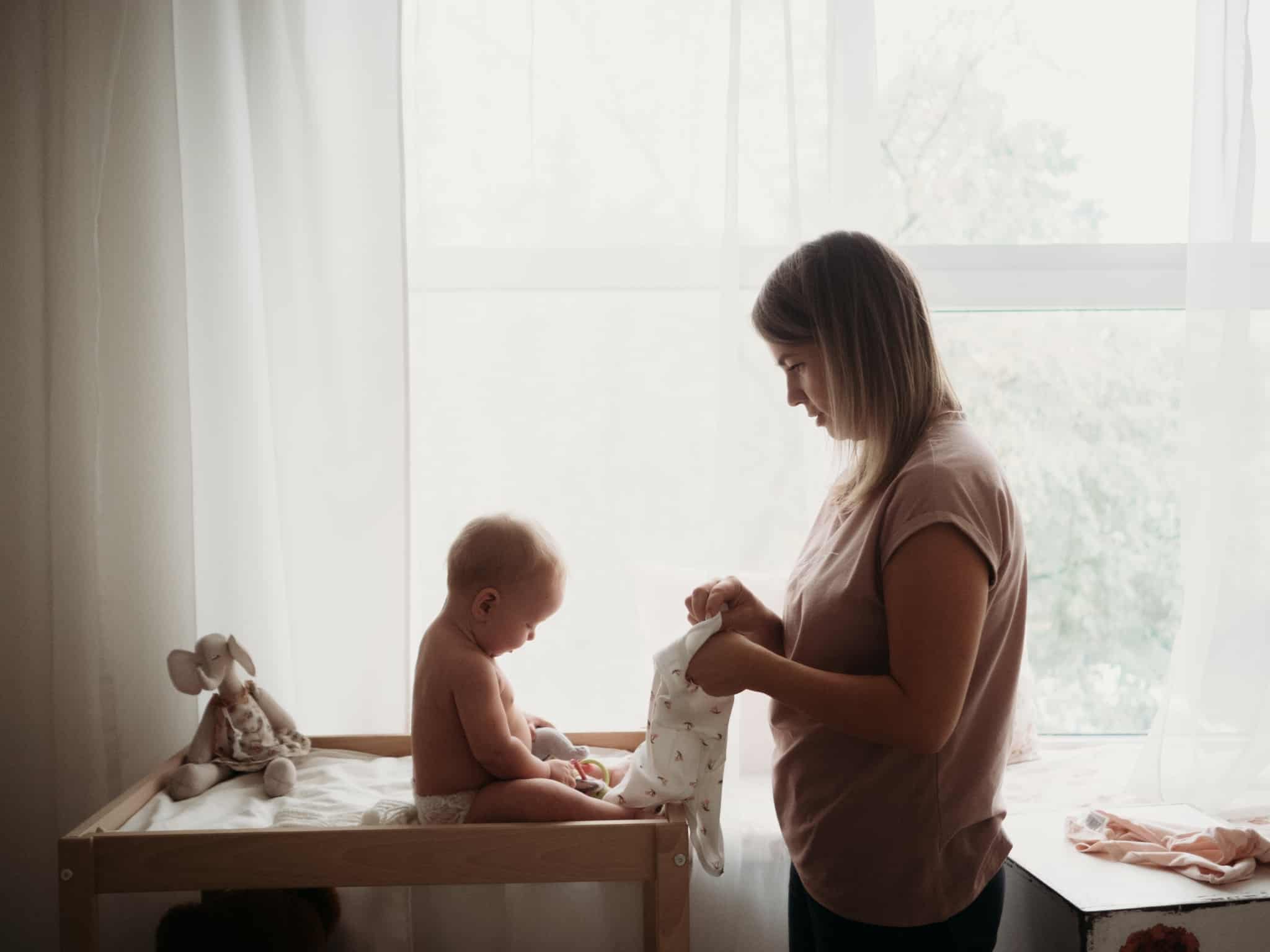 jovem mãe veste o seu bebé, no seu quarto, rodeado de todos os bens essenciais aos seus cuidados