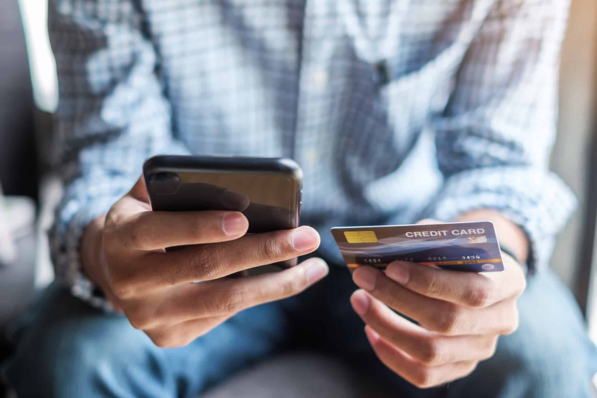 homem sentado com um telemóvel numa mão e um cartão de crédito na outra mão