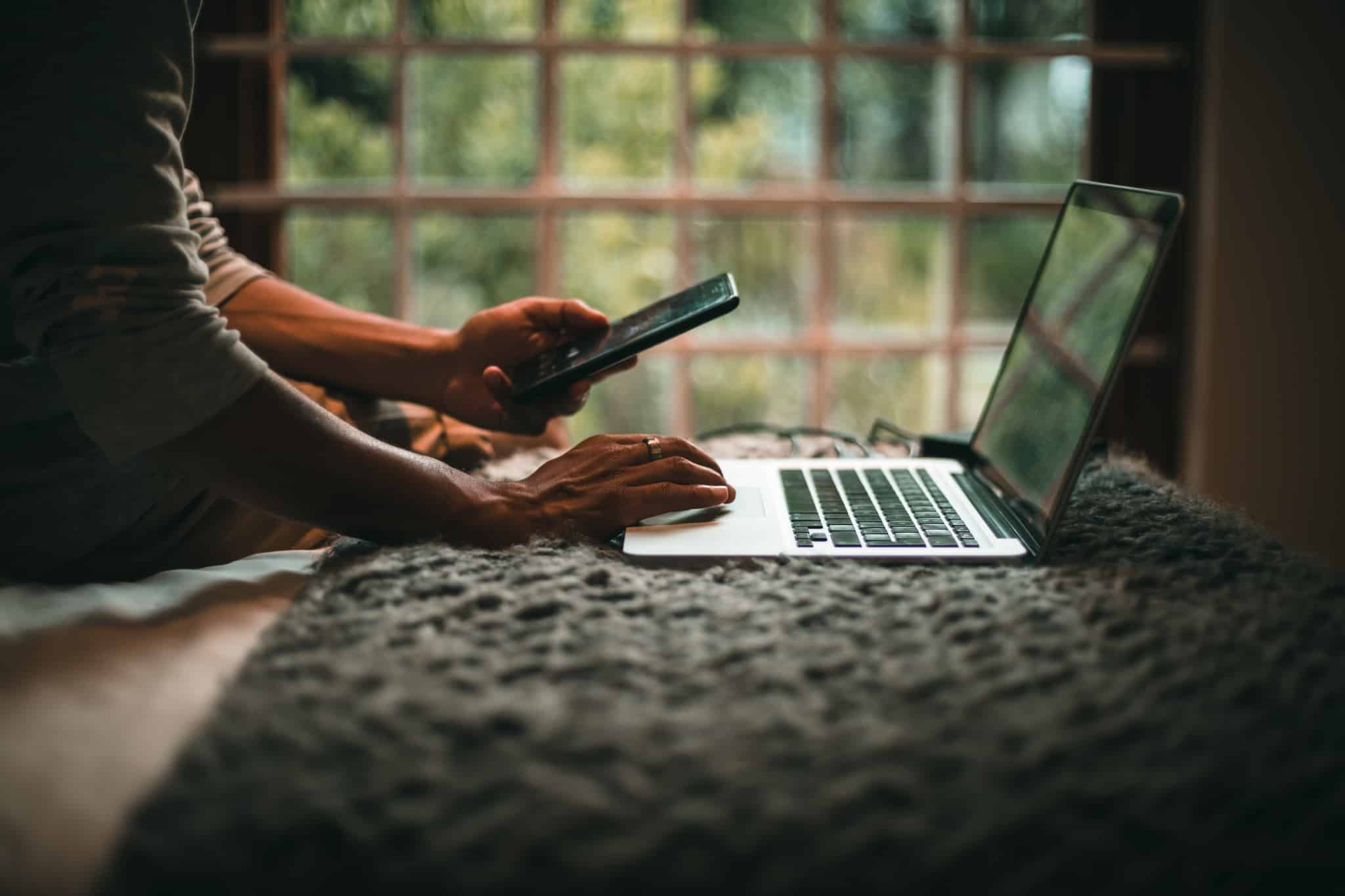 homem consulta o seu telemóvel enquanto trabalha em frente do seu computador. Ao fundo estão janelas antigas que precisam de serem substituídas