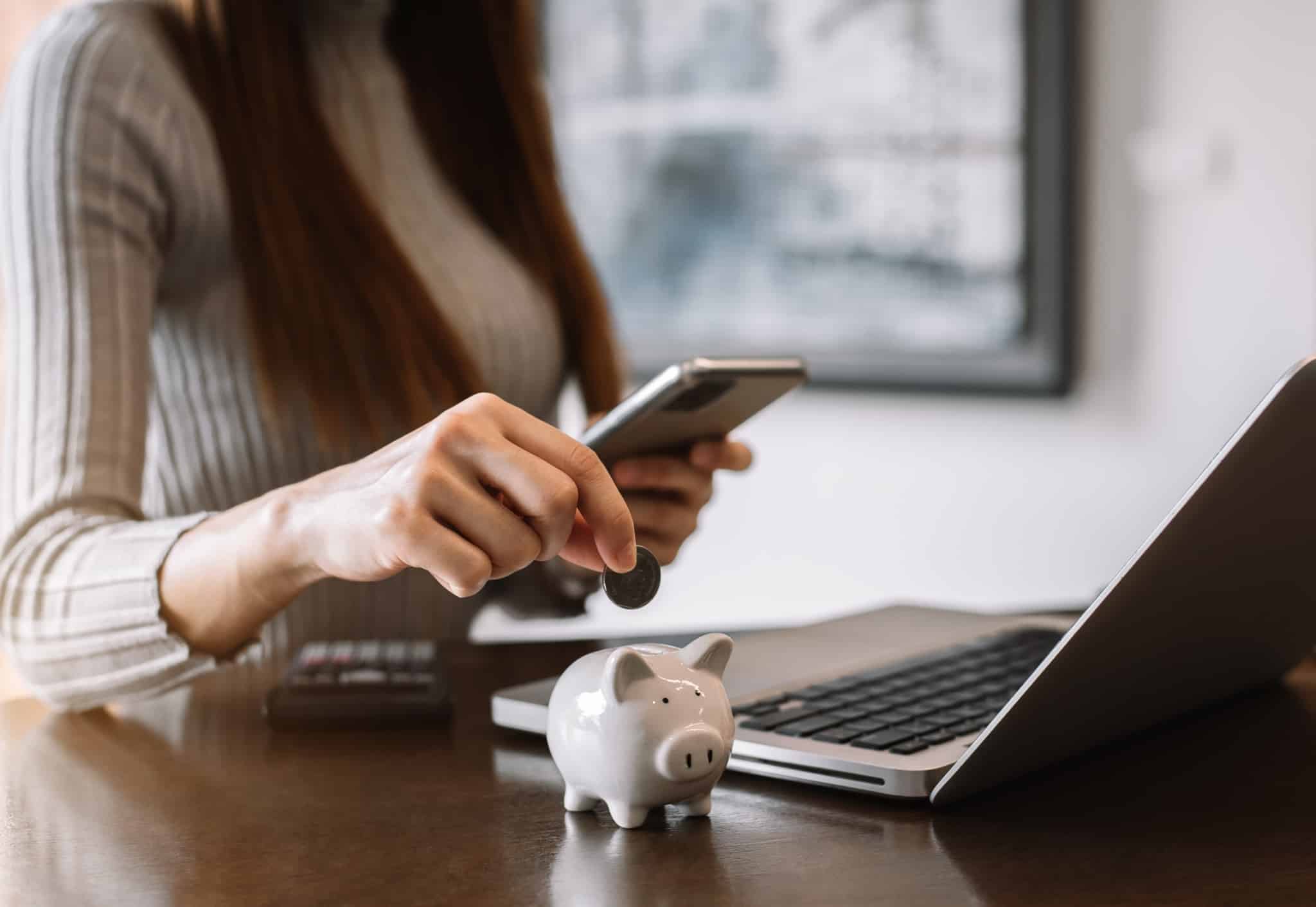 jovem mulher consulta telemóvel e juno ao seu porquinho mealheiro, usa o telemóvel e o computador portáti para planear as suas poupanças