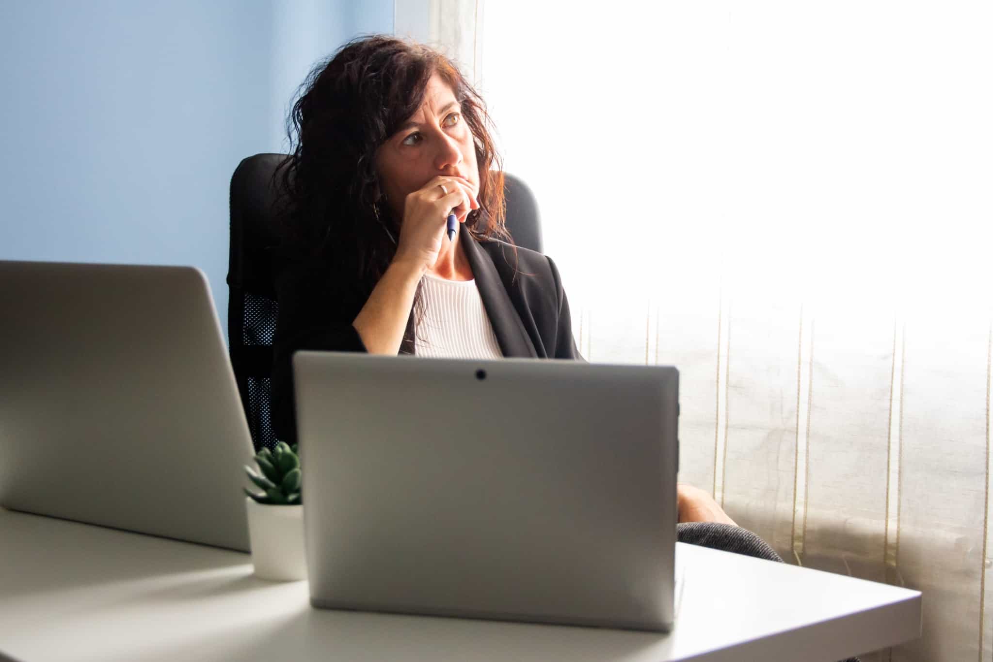 mulher sentada frente aos computadores de olhar triste e pensativa, como que a questionar-se ou a refletir