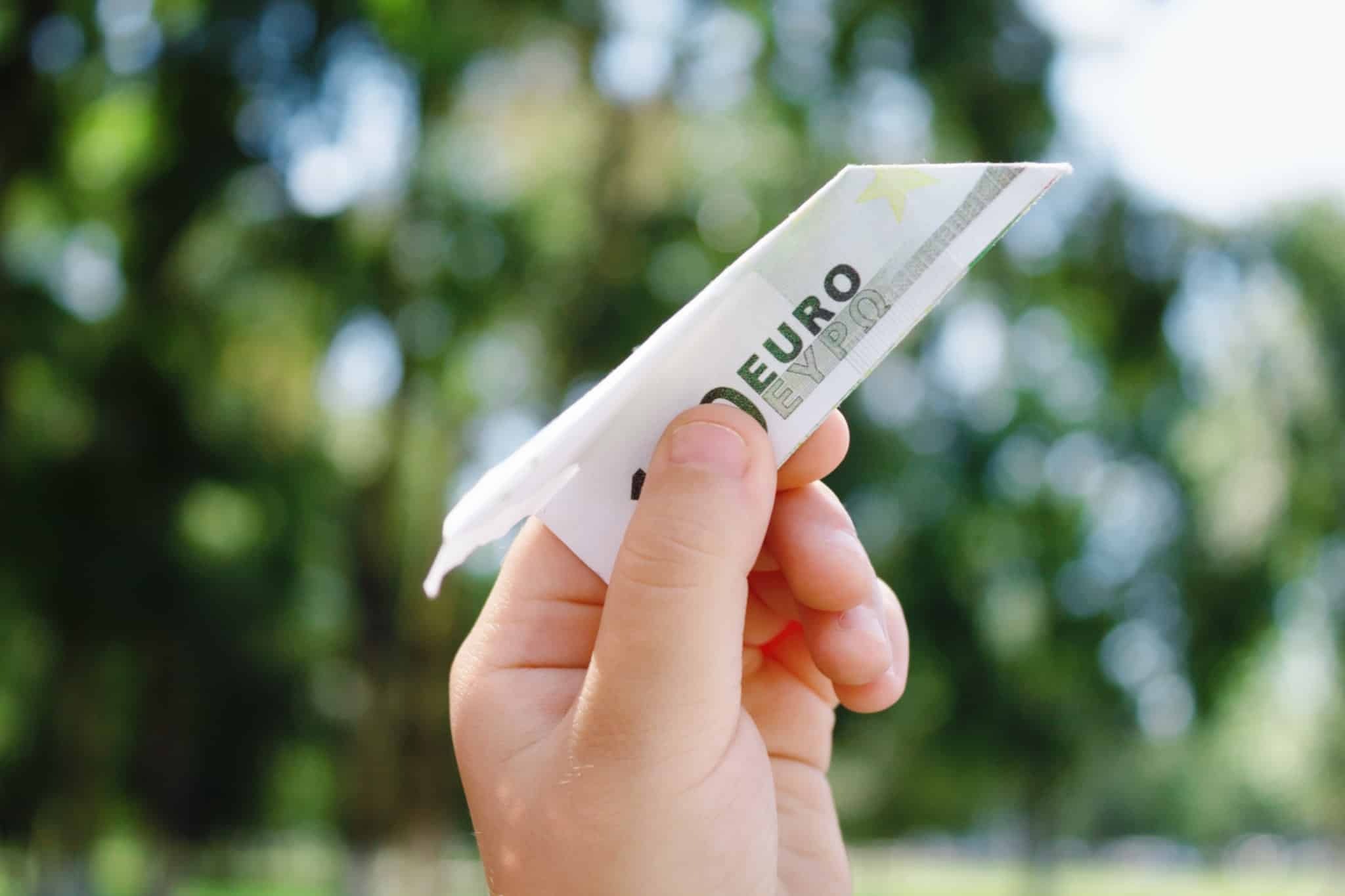 mão de ciança segura um avião de papel feito com uma nora de 5 euros que está pronto para se lançado ao ar