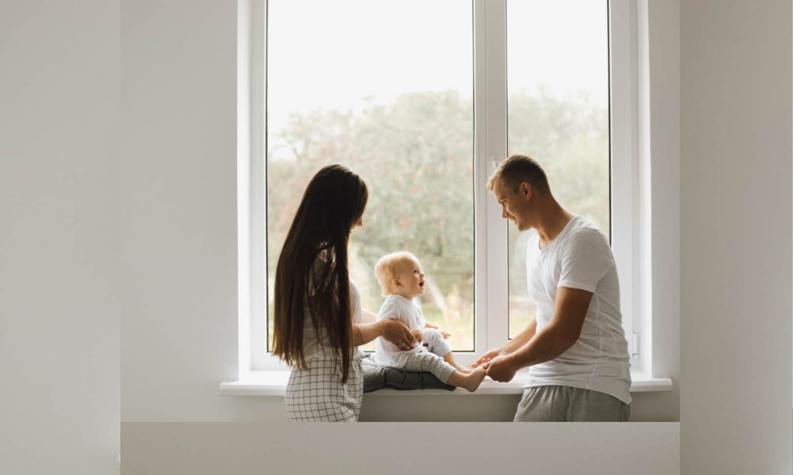 casal com um bebé junto a uma grande janela de sua casa, usufruindo de o subsídio de adoção