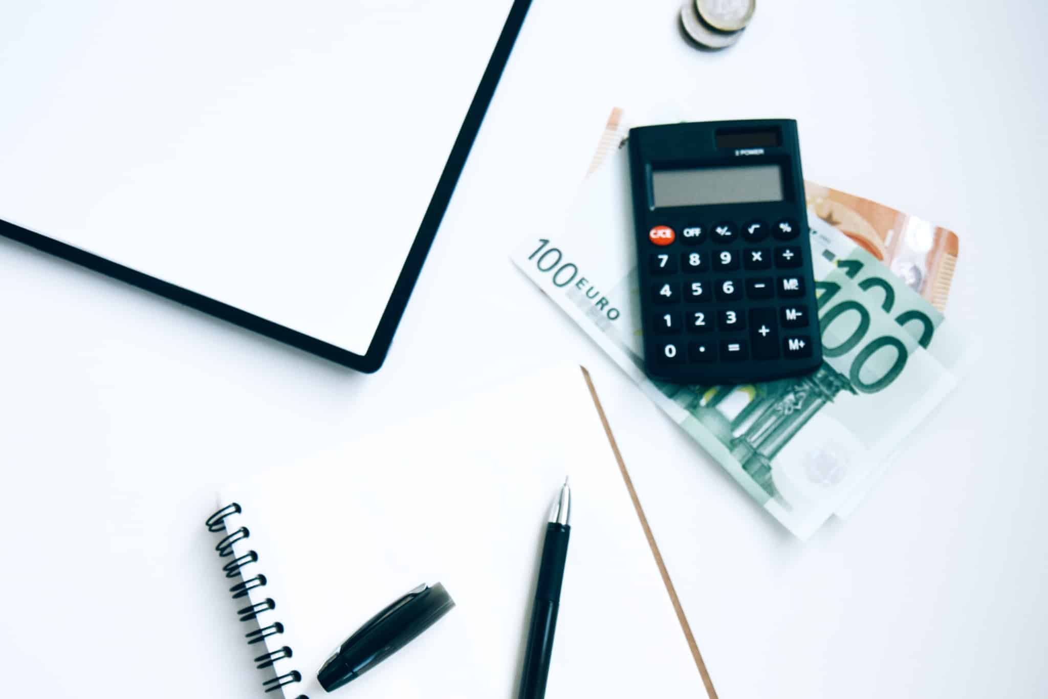 produção fotográfica com uma pequena calculadora preta em cima de algumas notas de 100 e 50 euros, ao lado de uma caderno em branco com uma caneta
