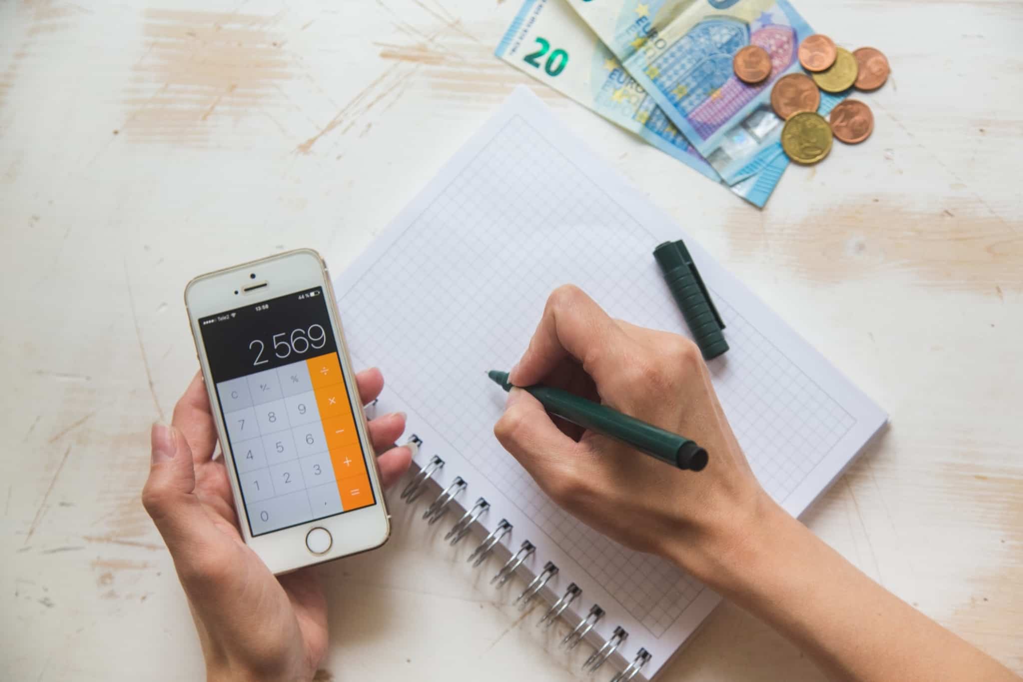 mãos de um jovem que seguram uma calculadora e uma ce nta, com os quais fazem contas tamb~em num caderno, enquanto na mesa se vê algumas notas e moedas de euro