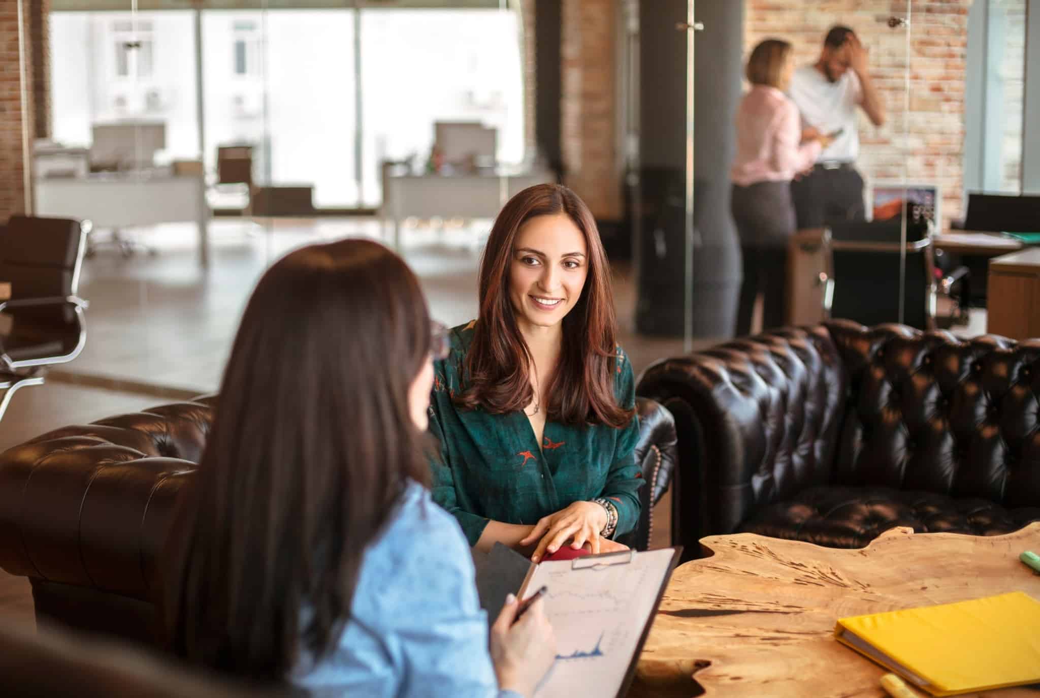 ambiente de escritório com duas jovens mulheres em conversa, a analisar dodumentos, e no fundo, dois colegas trocam informações