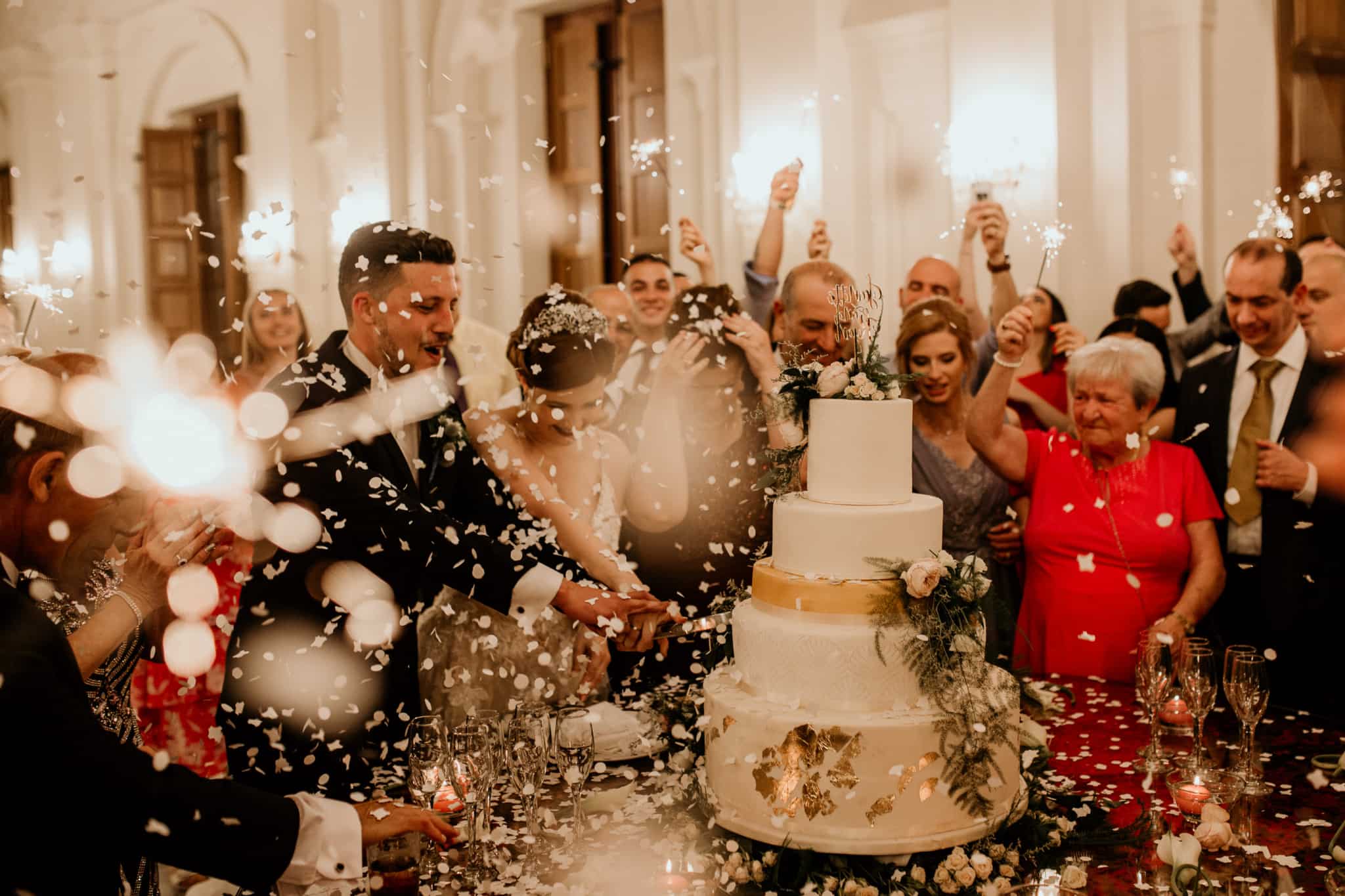 casamento vivido em grande ambiente de festa com os noivos rodeados pela família e amigos que brindam enquanto eles se preparam para cortar o bolo
