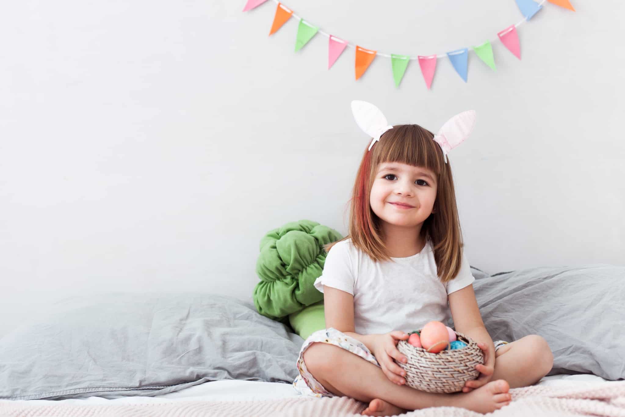 menina com bandolete de orelhas de coelho, enmtre uma decoração festiva, mostra um cesto cheio de ovos da Páscoa que decorou durante as férias da Páscoa