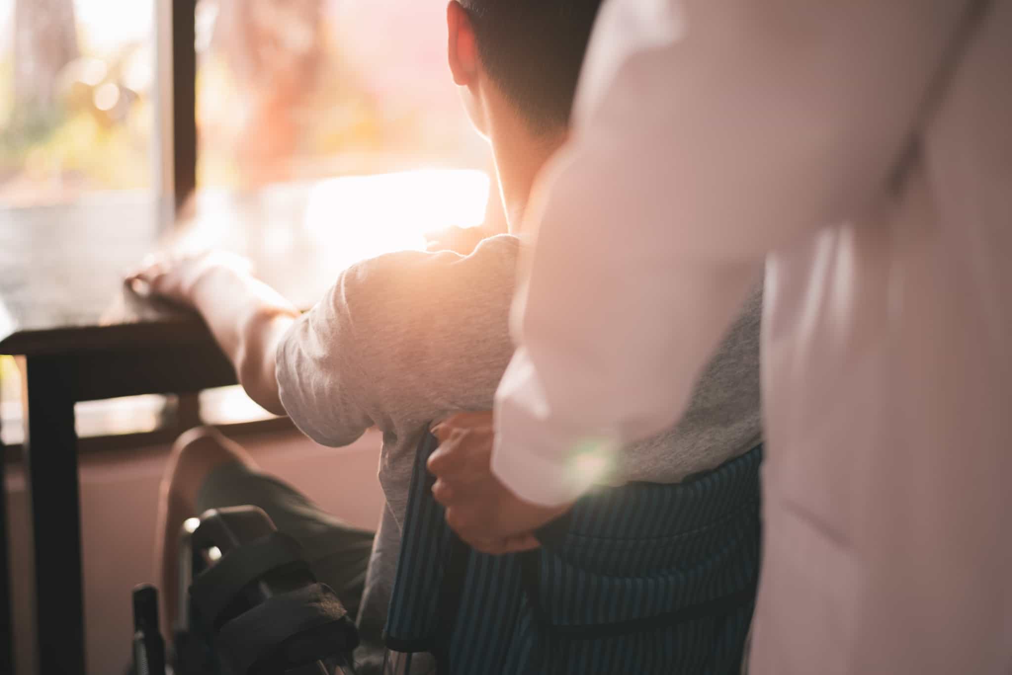 pessoa conduz jovem em cadeira até mesa, estando totalmente depende e a receber subsídio por assistência de terceira pessoa