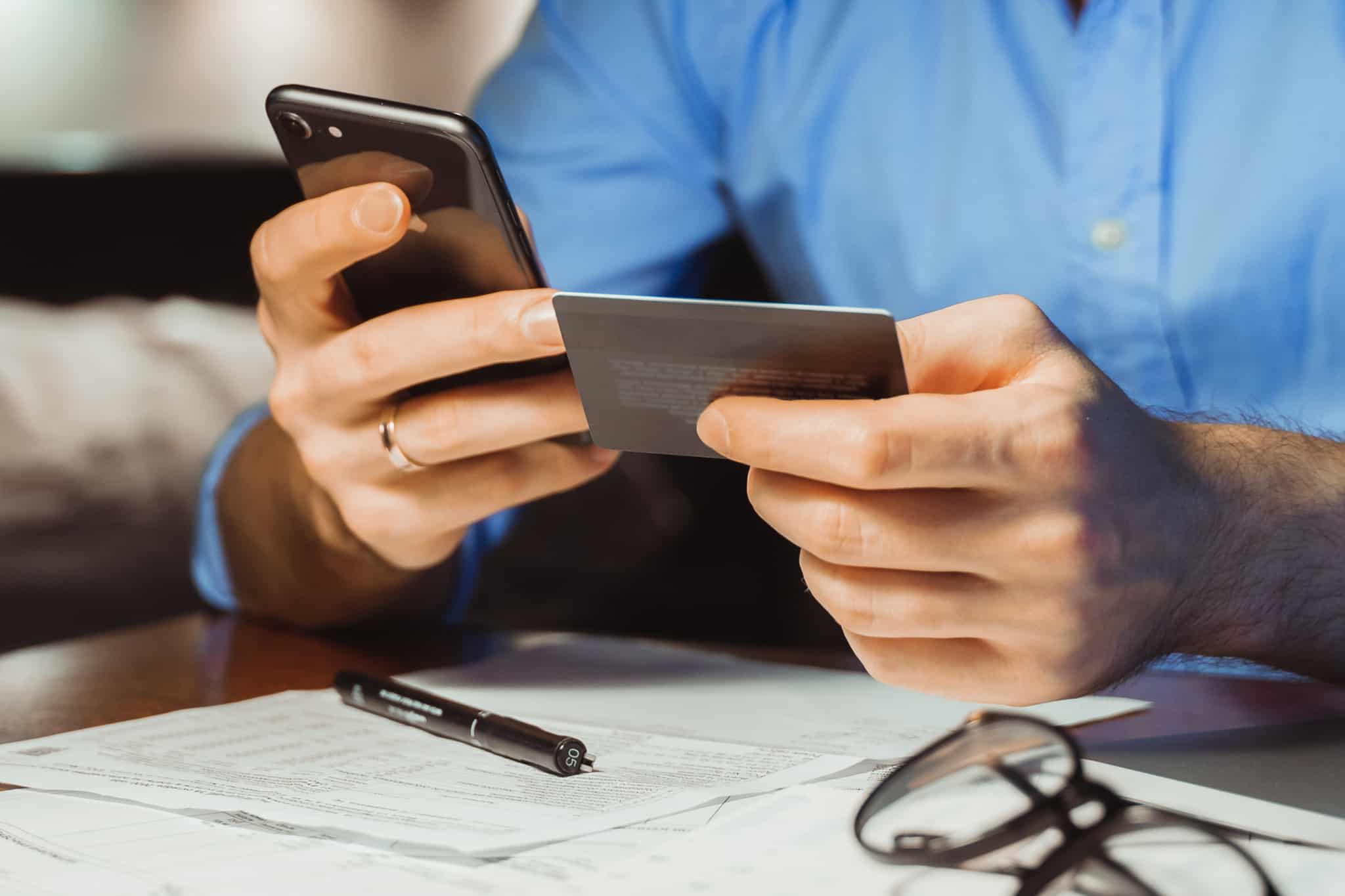 Homem trabalhador independente a segurar num telemóvel e cartão bancário, com faturas irs em cima da mesa