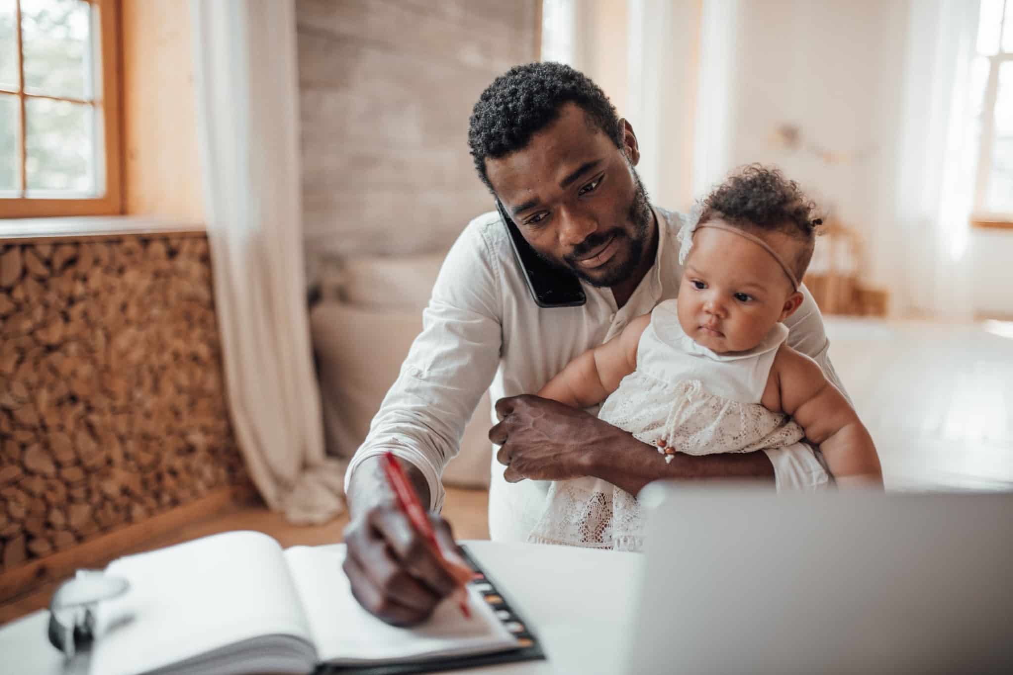 Imagem de um pai com a filha pequena ao colo, à mesa, com um pc portátil, enquanto escreve num caderno - numa alusão ao IRS para pais divorciados