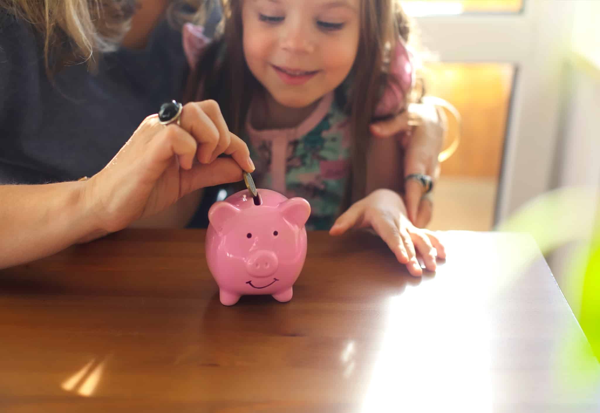 mãe com a filha pequena ao colo, em casa e à mesa, ajuda-a a gostar de finanças pessoais com o exemplo da poupança em porquinhos mealheiros