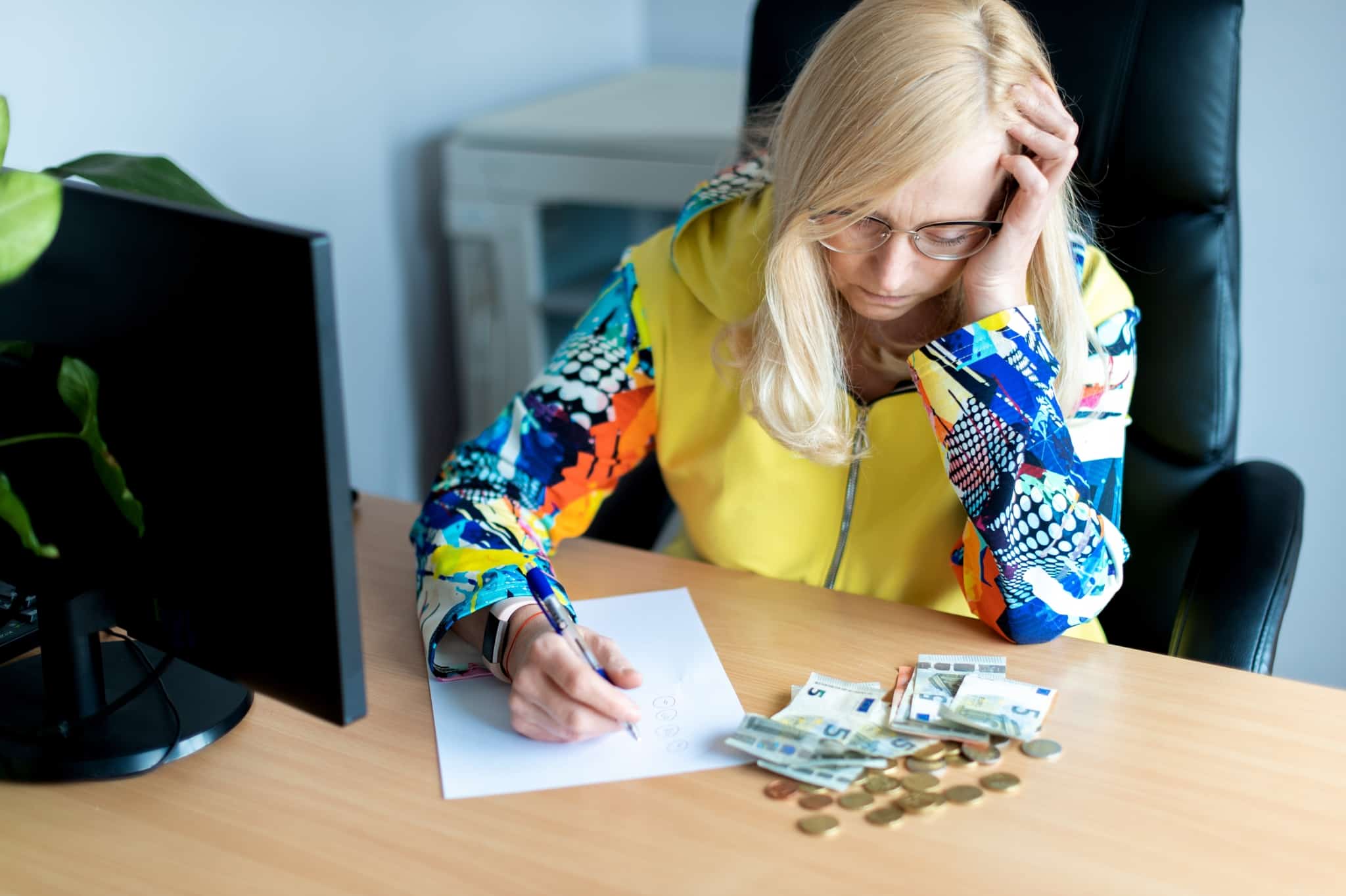 mulher, de meia idade, com lápis e folha de papel, sentada à mesa onde tem espalhado moedas e notas de cinco euros, tenta fazer o seu orçamento contando apenas com rendimentos irregulares