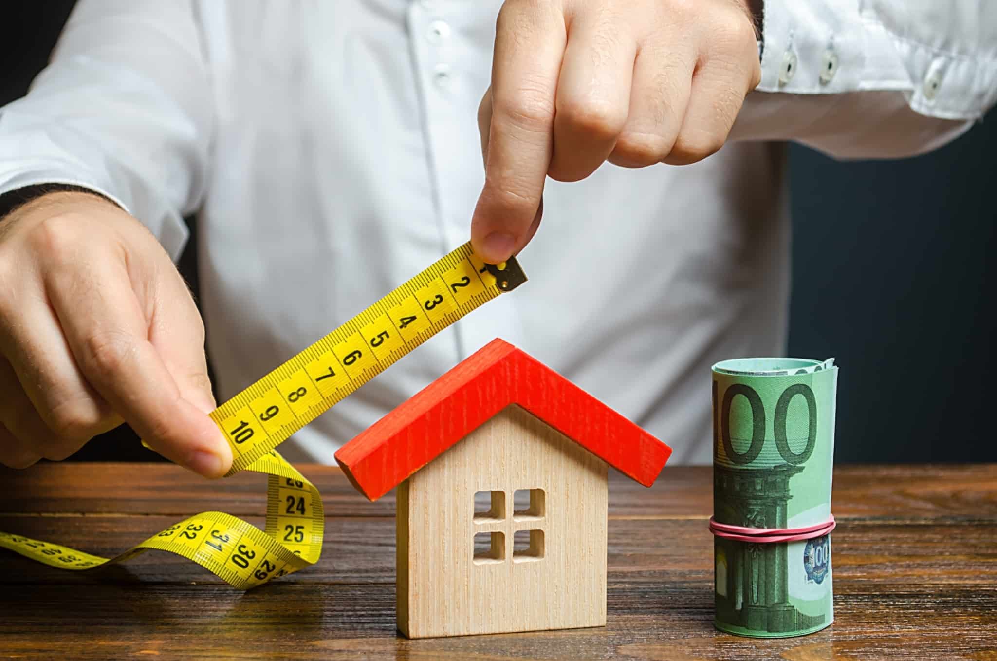 um homem a medir o telhado de uma casa de madeira com um rolo de notas ao lado, servindo como referência a subida do valor dos imóveis e também aos mitos sobre o crédito habitação