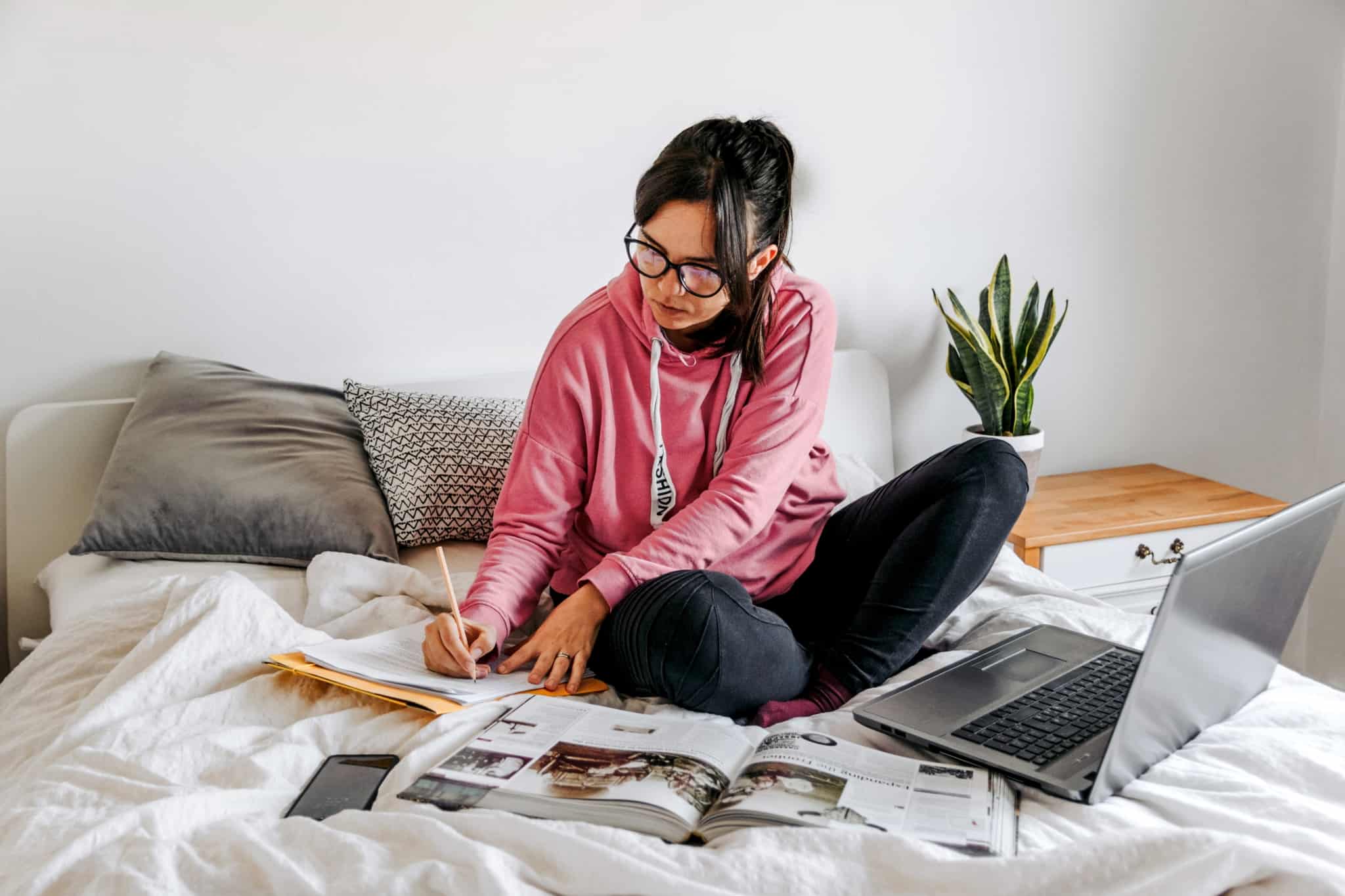 jovem mulher estudante, rodeada de livros e com o computador portátil, sentada na cama, procura formas de ajudar a pagar o curso
