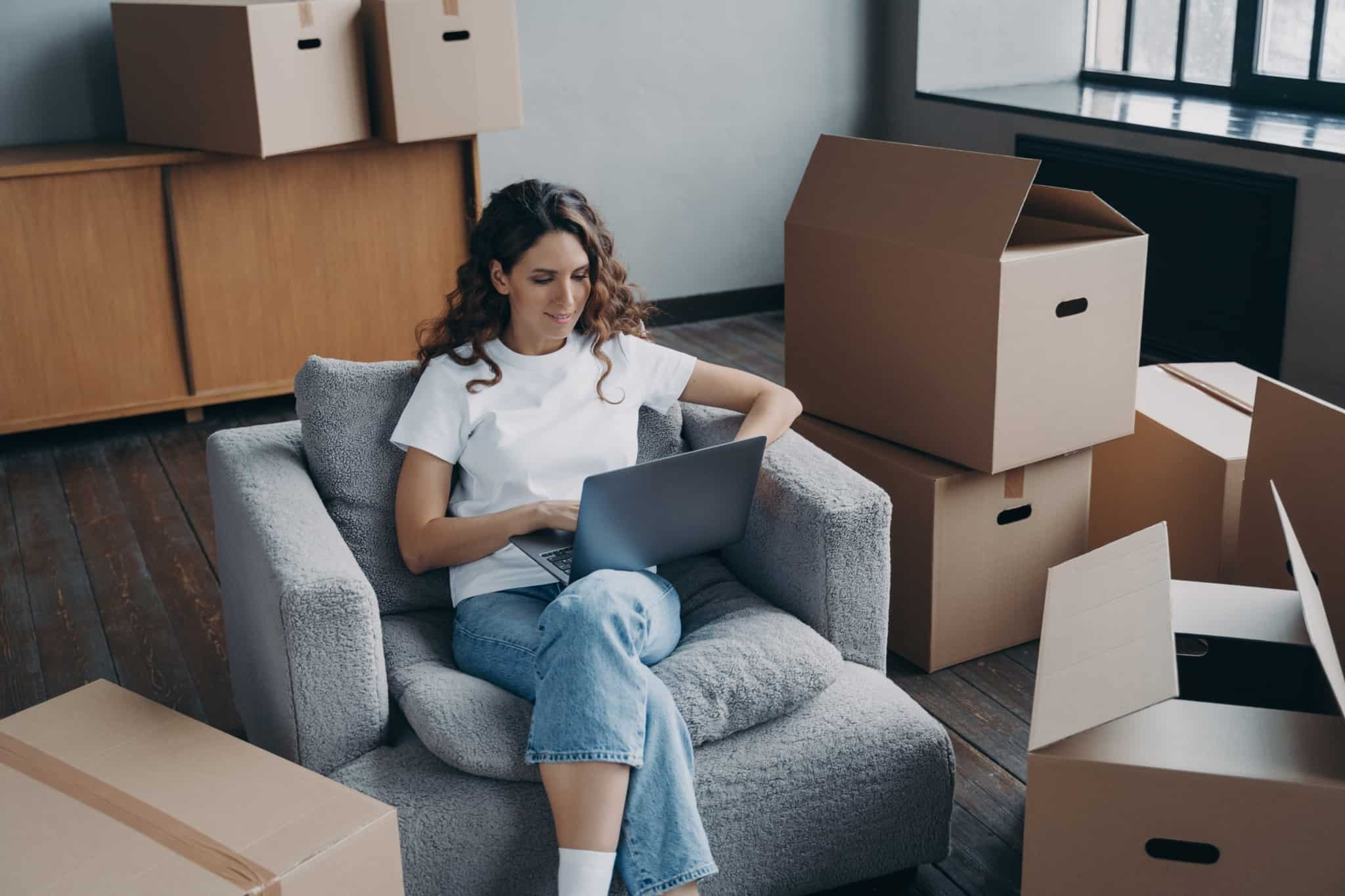 jovem mulher, entre caixas de mudança e ao computador portátil, sentada no sofá planeia mudar de cidade por um novo emprego