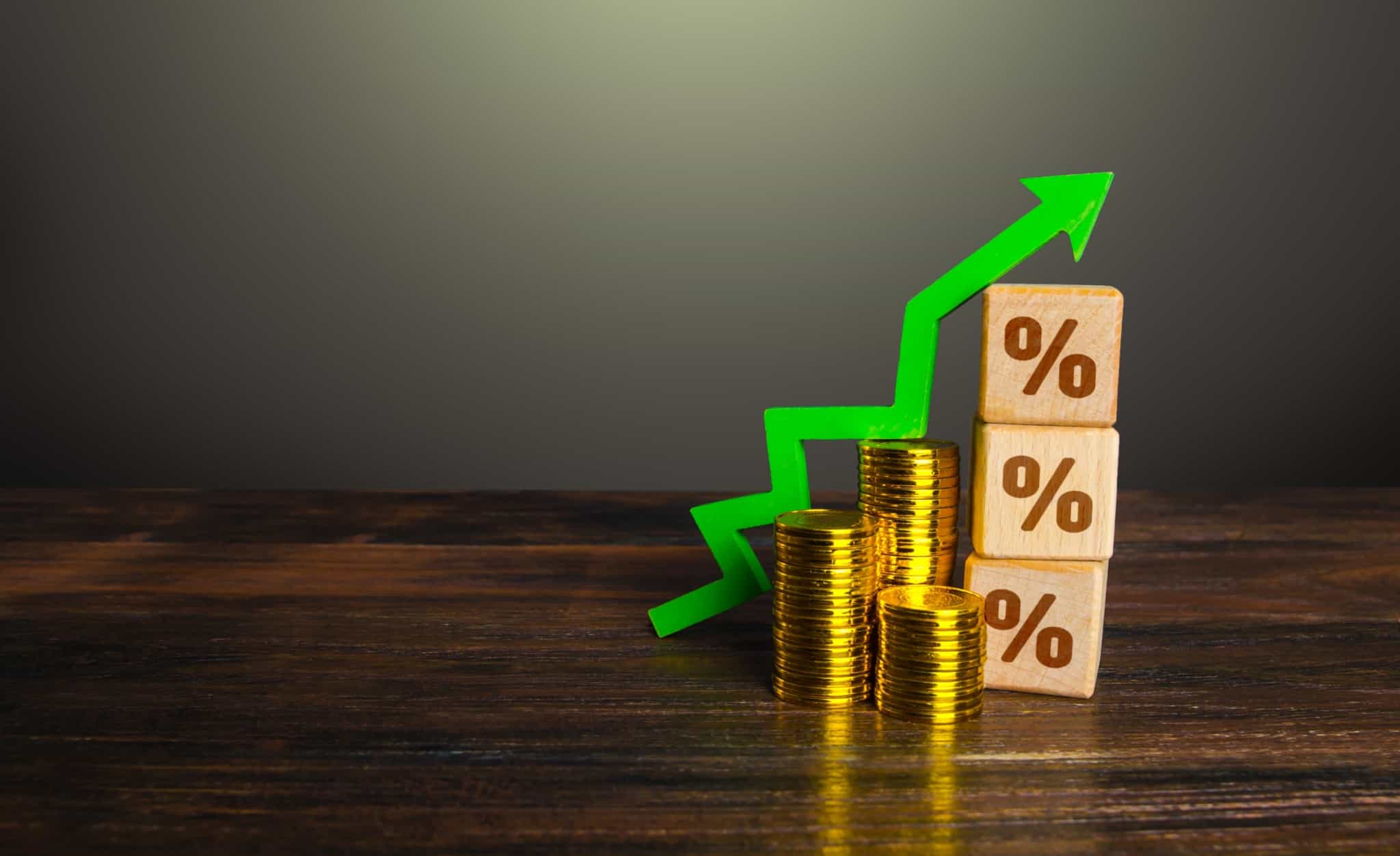 produção fotográfica, com fundo escuro, mostra pequenas pilhas de moedas, junto a três cubos com o símbolo da percentagem e com um uma seta verde ascendente, remetendo assim para a subida das taxas de juro