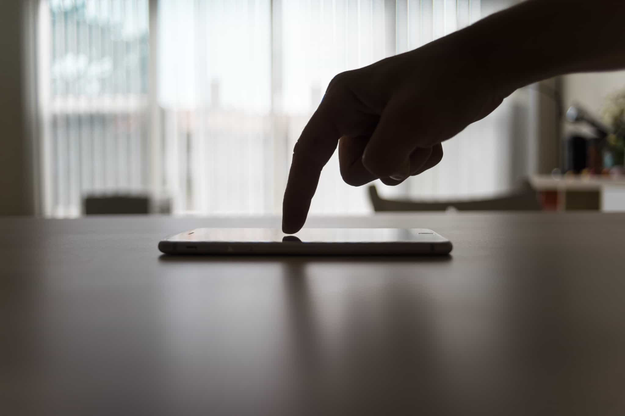numa imagem em contraluz, um dedo percorre o ecran de um telemóbel, em cima de uma mesa, enquanto acede á conta bancária no telemóvel