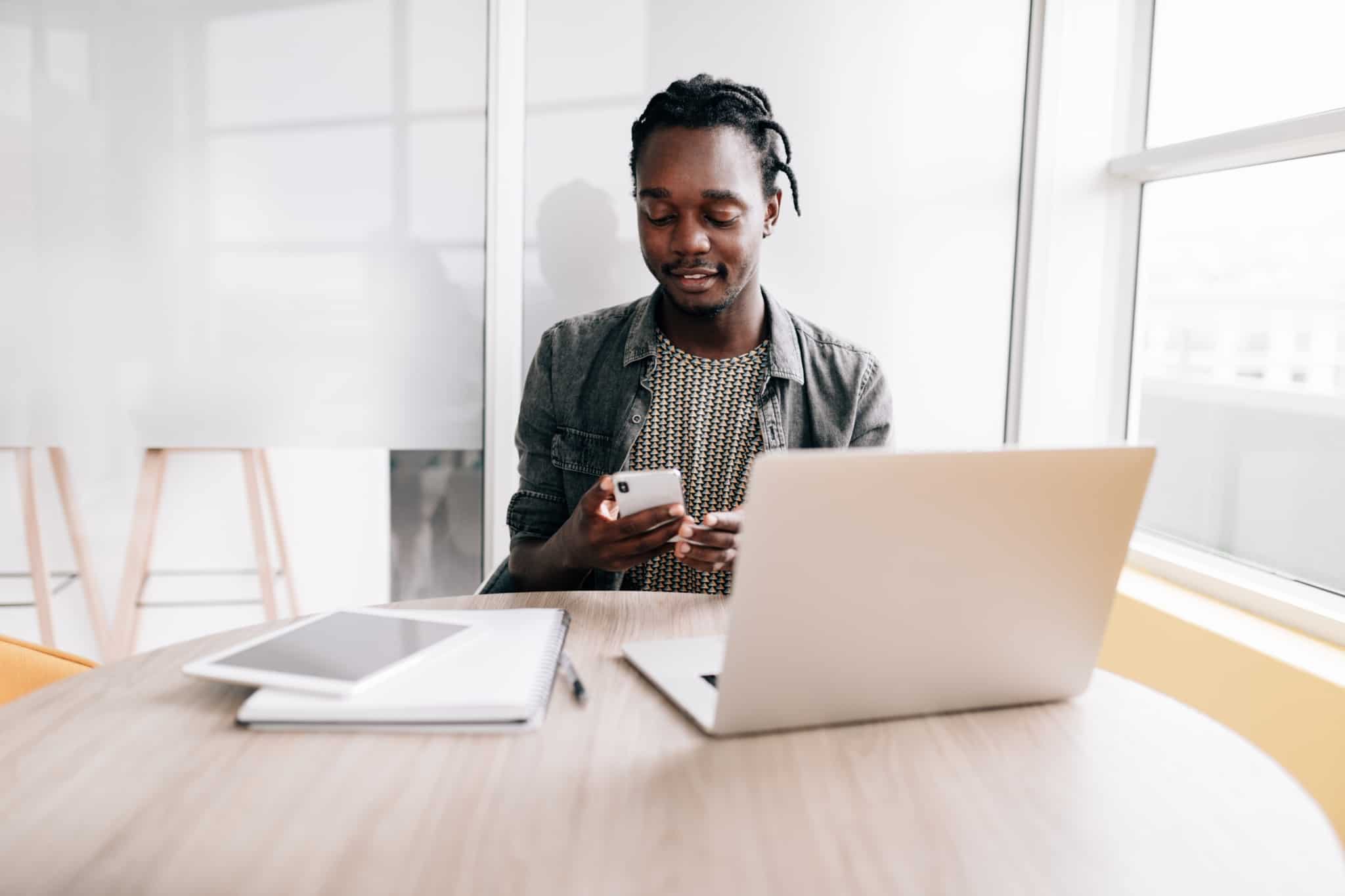 Rapaz sentado à secretária com computador, telemóvel e caderno em teletrabalho