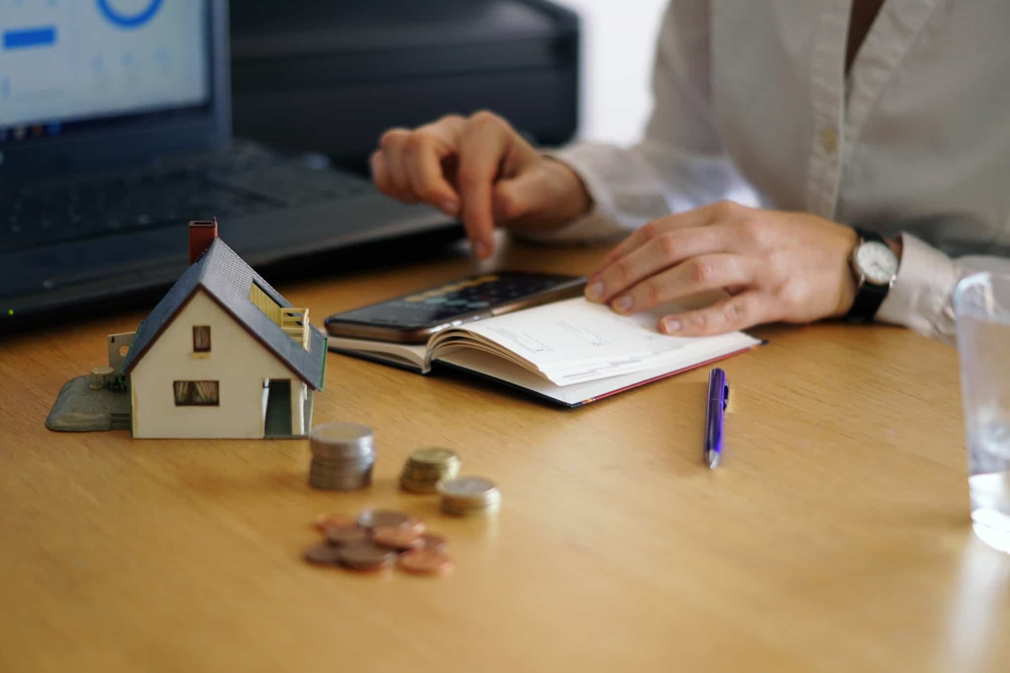 jovem rapaz a fazer contas no caderno e com uma calculadora, sentado à mesa com uma pequena maqueta de uma casa e algumas pilhas de moedas, analisa propostas de crédito habitação