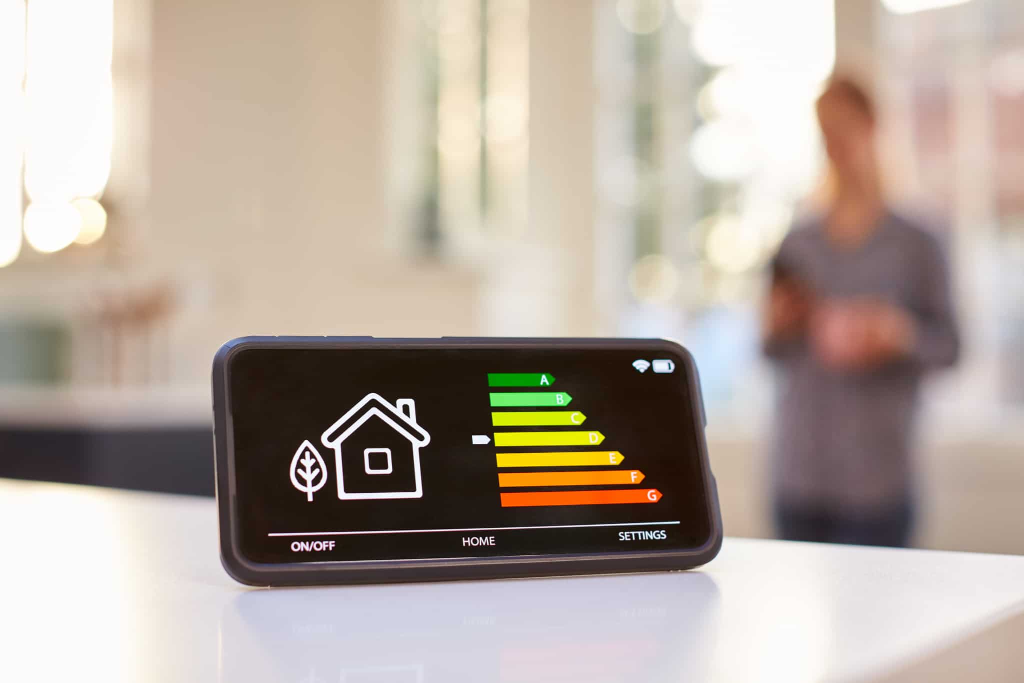 ecran de um smartphone mostra etiqueta energética de uma casa, enquanto uma pessoa conytrola essa app na sala, iliustrando o que pode ser a gestão da eficiência da energia e da água