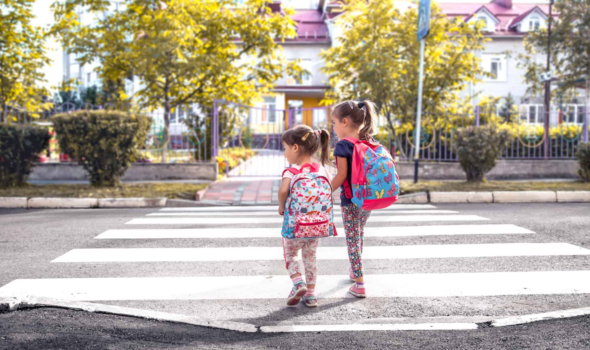 duas meninas, de mochilas às costas e de mãos dadas, atravessam na passadeira frente ao portão da escola, rementendo para as questões de segurança e do acidente na escola