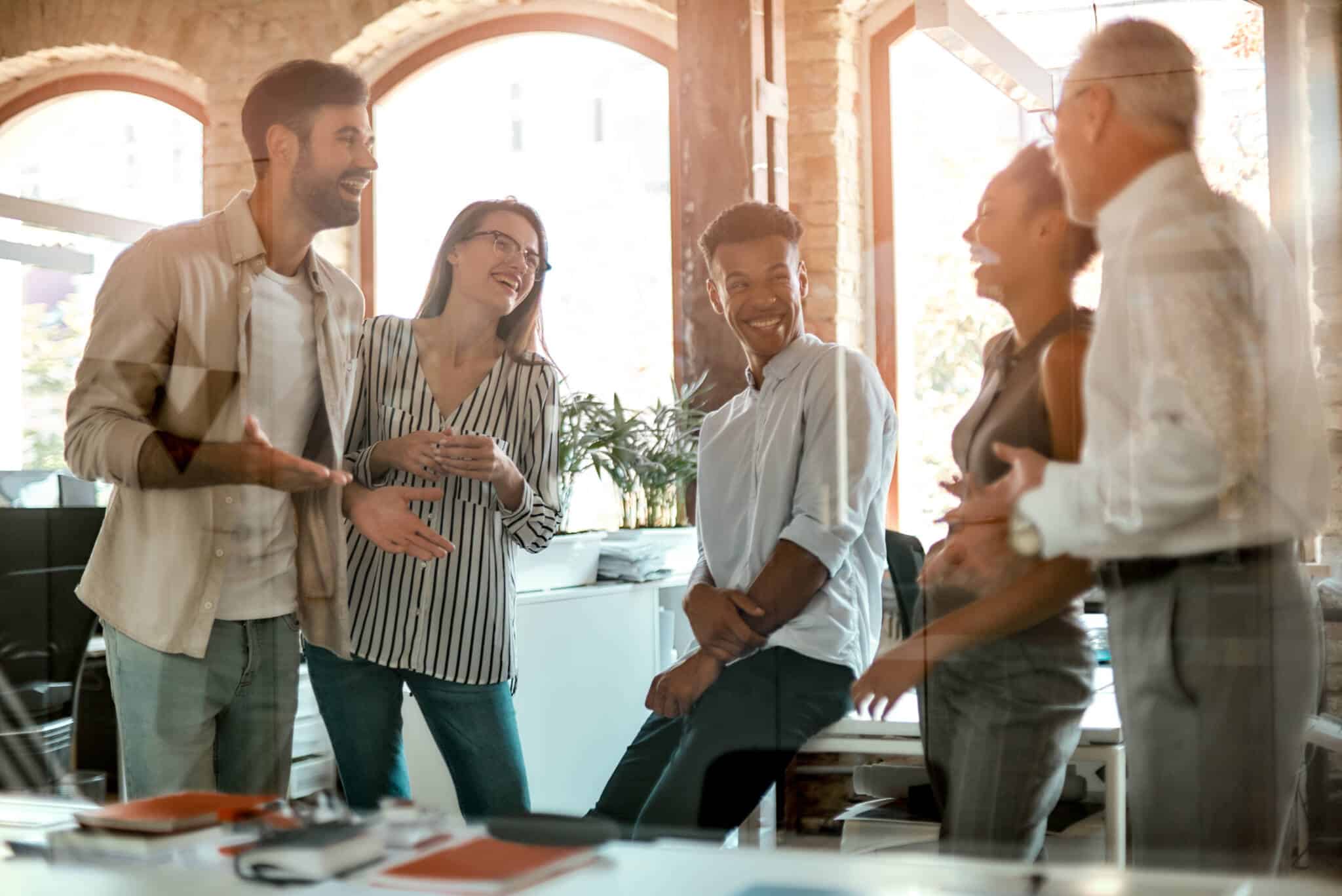 momento descontraído e gargalhada partilhada pr um grupo de colegas, homens e mulheres, no seu ambiente de trabalho, remetendo para a importância, caso queira mudar de emprego, ter de fazer contas ao salário emocional