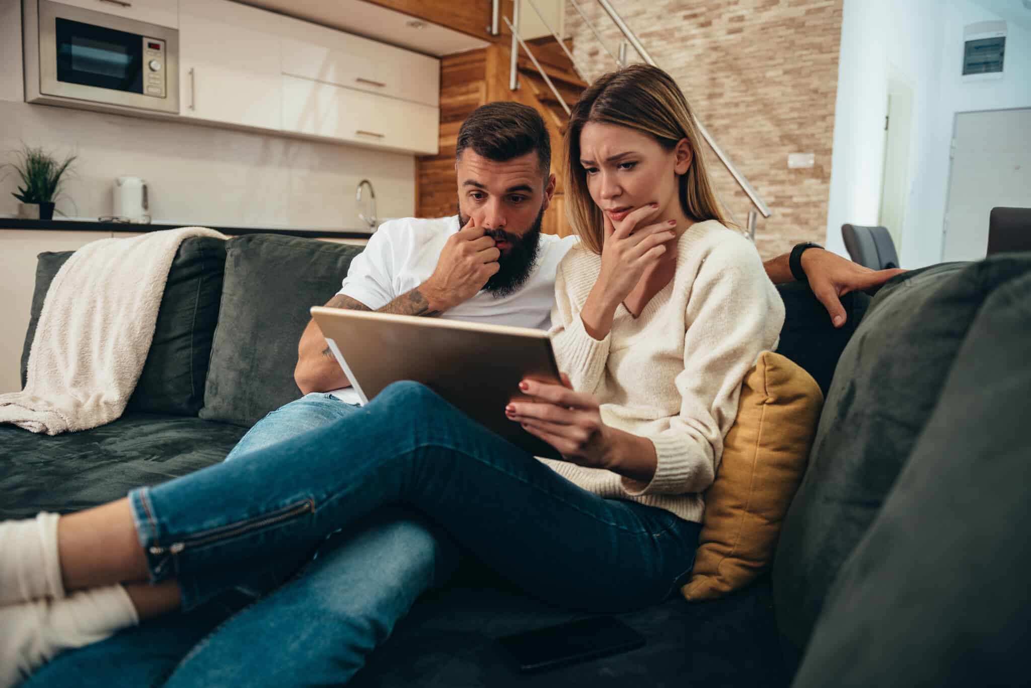 jovem casal, sentados no sofé da sua sala de estar, consulta no tablet o seu orçamento de férias, constatando, com um ar preocupado, que tiveram gastos extra que vão ter de compensar