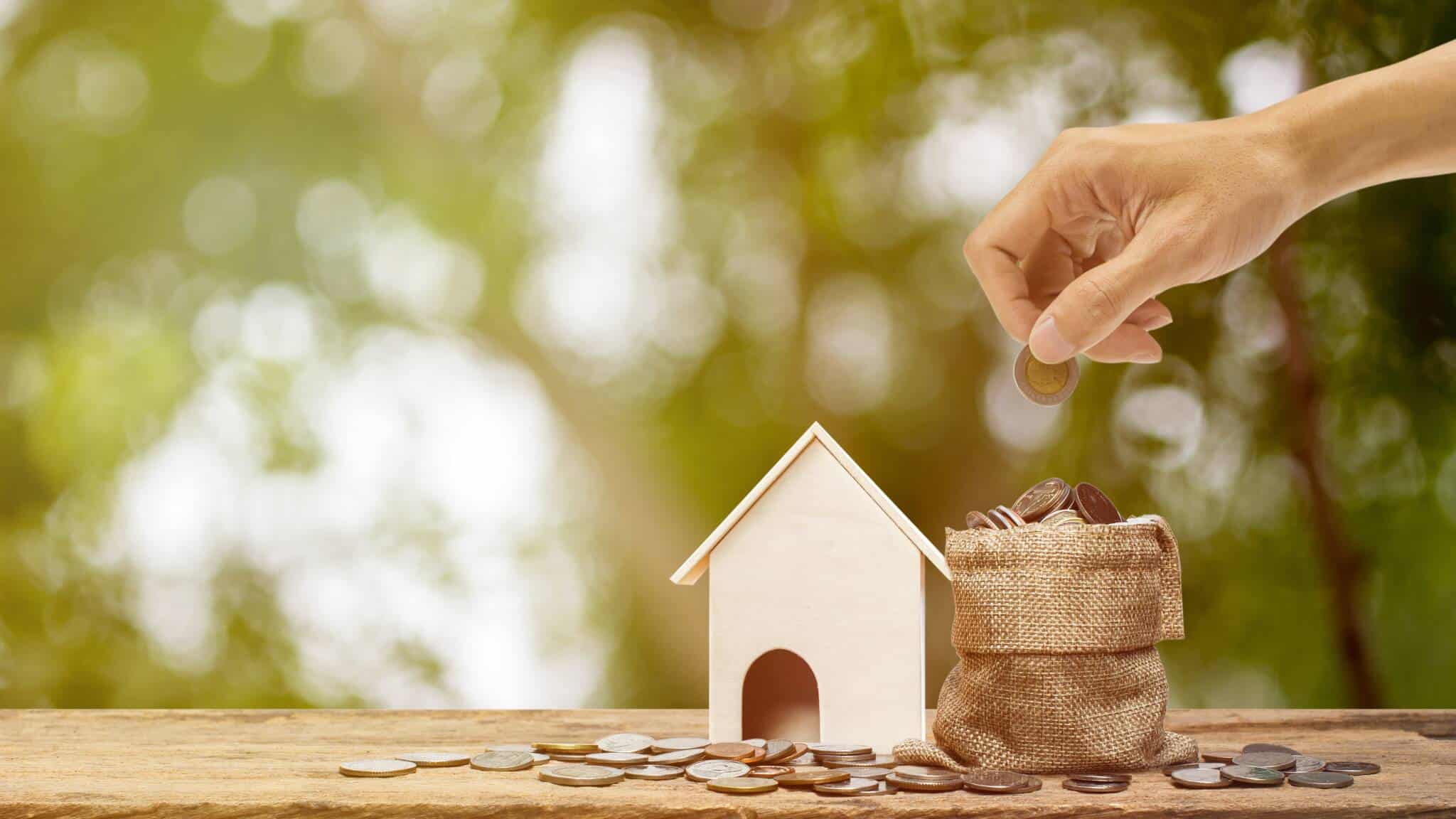Casa de madeira com saco ao lado com moedas e mão a colocar moeda no saco