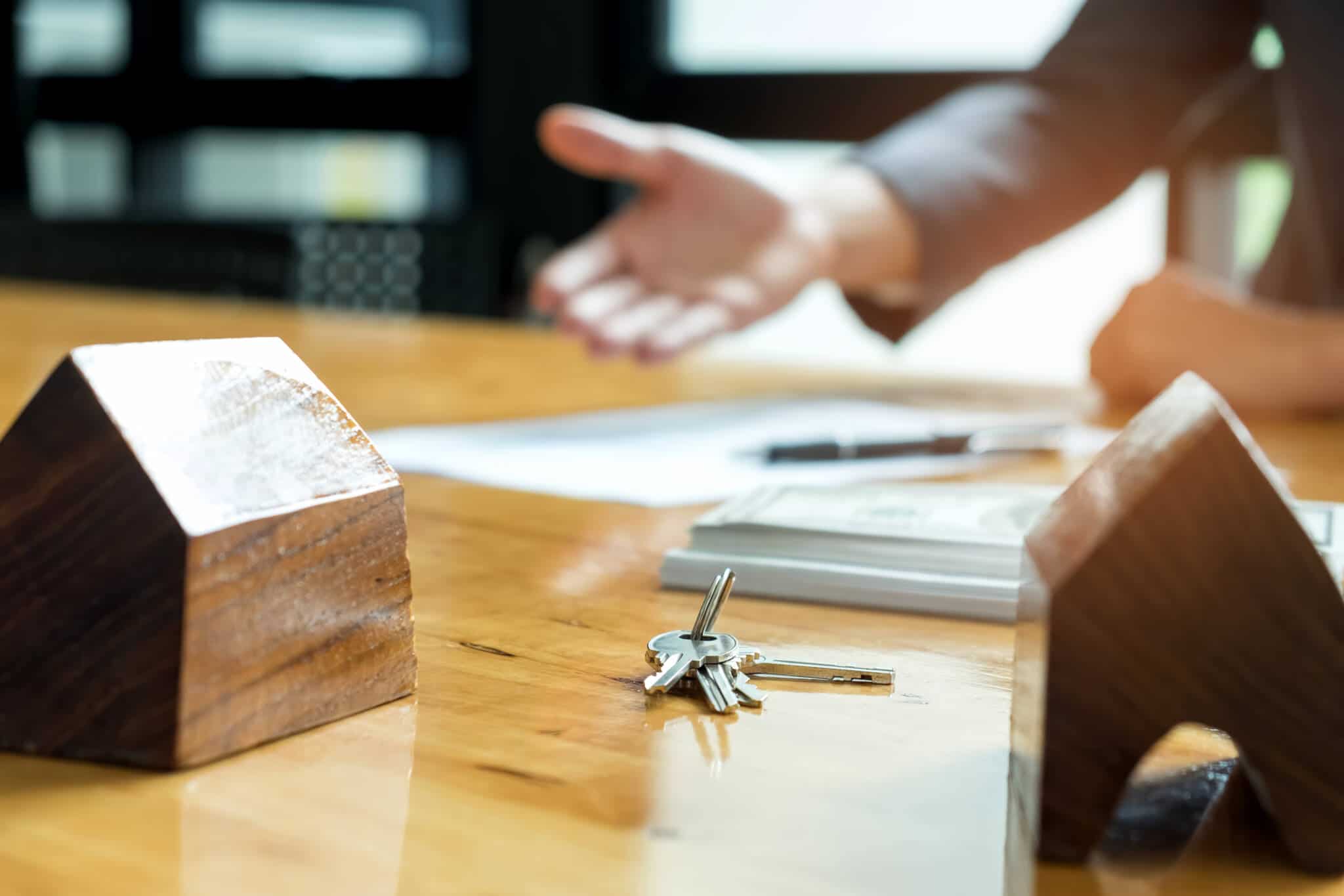 em cima de uma mesa estão duas maquetas de casas, em madeira, junto a papeis e umas chaves de casa, num conjunto exposto por um jovem homem que explica o que fazer para vender imóvel herdado