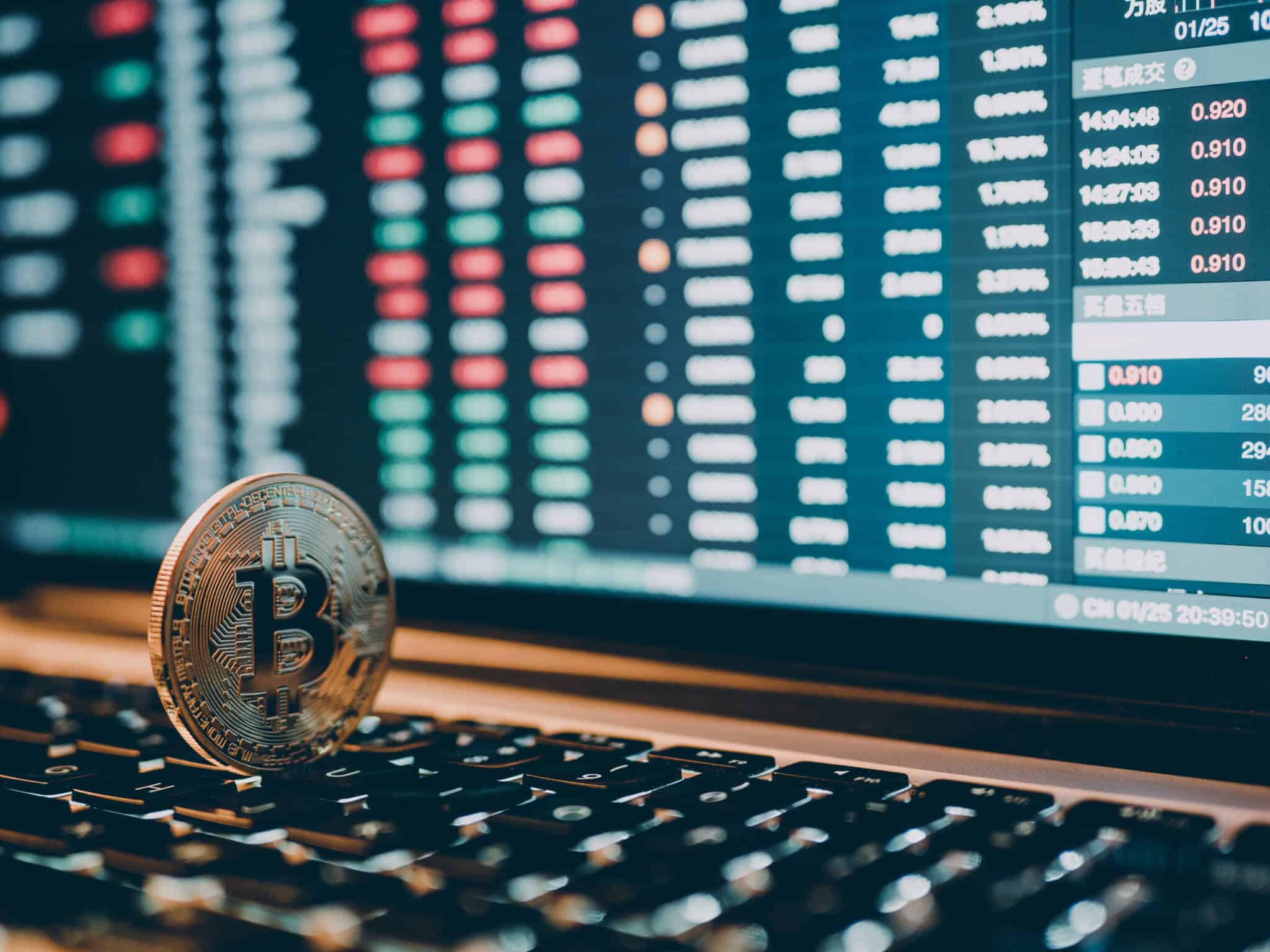 Imagem de uma criptomoeda sem cima de um teclado e de um monitor com gráficos numa alusão ao mercado de criptoativos