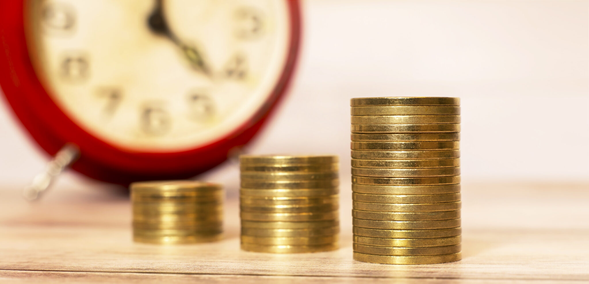 Poupanças e inflação ilustradas por três pilhas em crescendo de moedas sob um fundo desfocado de um relógio que marca o tempoe remete para um plano financeiro que as famílias devem ter