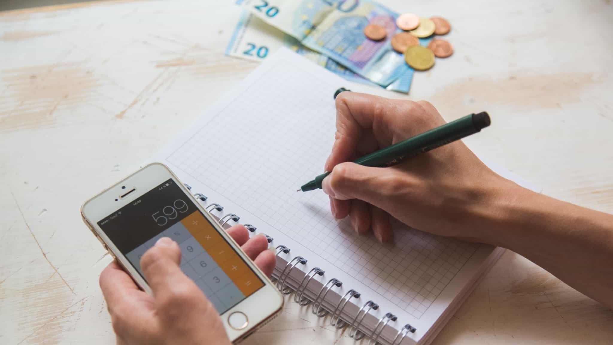numa mesa, com algumas notas de euro e moedas, vê-e apenas a mão de quem está a usar a calculadora e um caderno e caneta para afzer contas ao seu orçamento