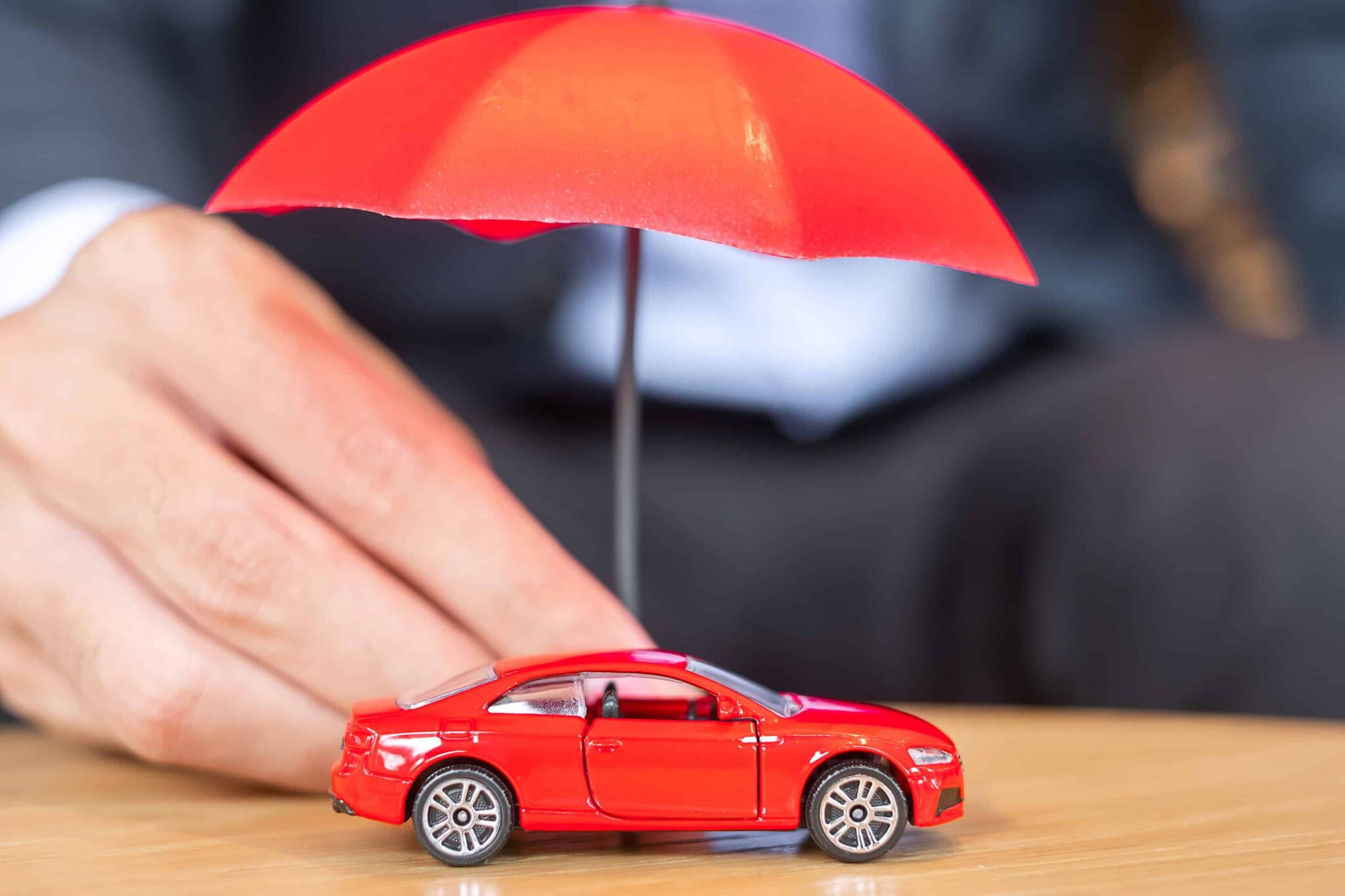 mão de homem a segurar um chapeu de chuva em miniatura, que está a cobrir um automóvel tambbém miniatura, ambos vermelhos, representando a possibilidade de verificar seguro pela matrícula