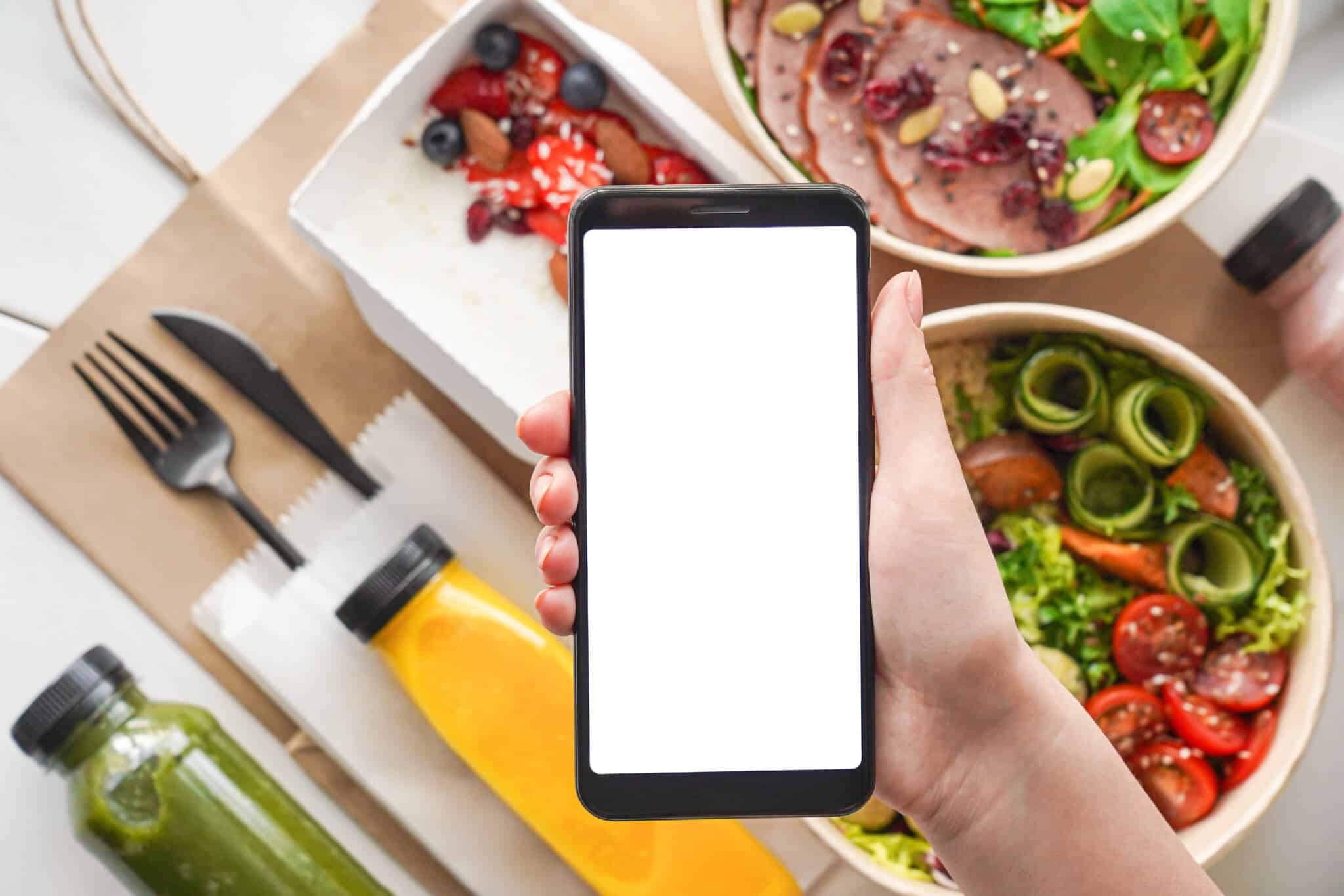 mão de jovem mulher segura telemóvel consultando aplicações para conseguir refeições mais baratas, sob um fundo de vários pratos com alimentos diveross e muito coloridos