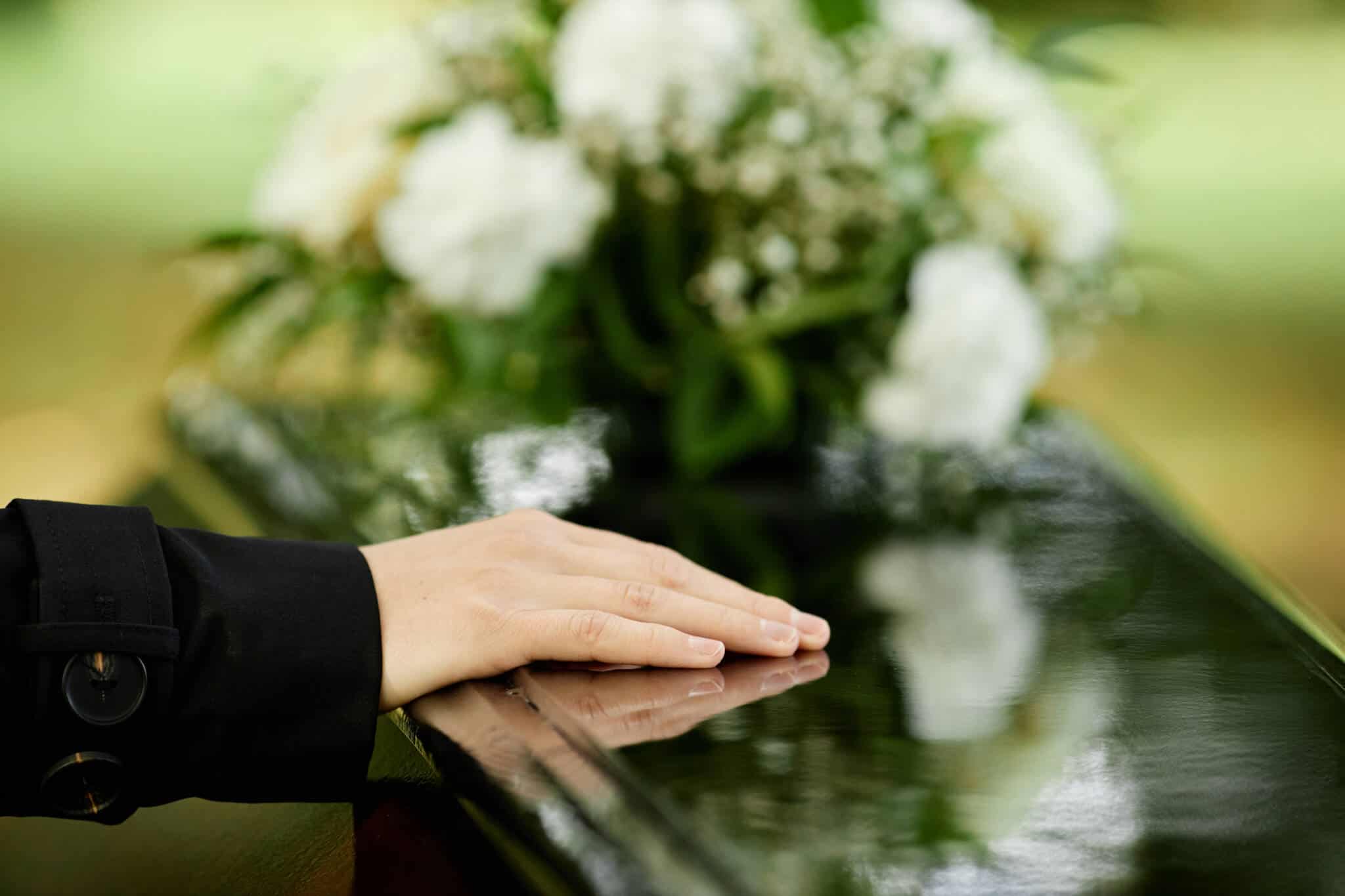 mão de jovem mulher repousa sob um caixão de madeira escura, com um ramo de flores desfocado em fundo, ilustrando o subsídio de funeral