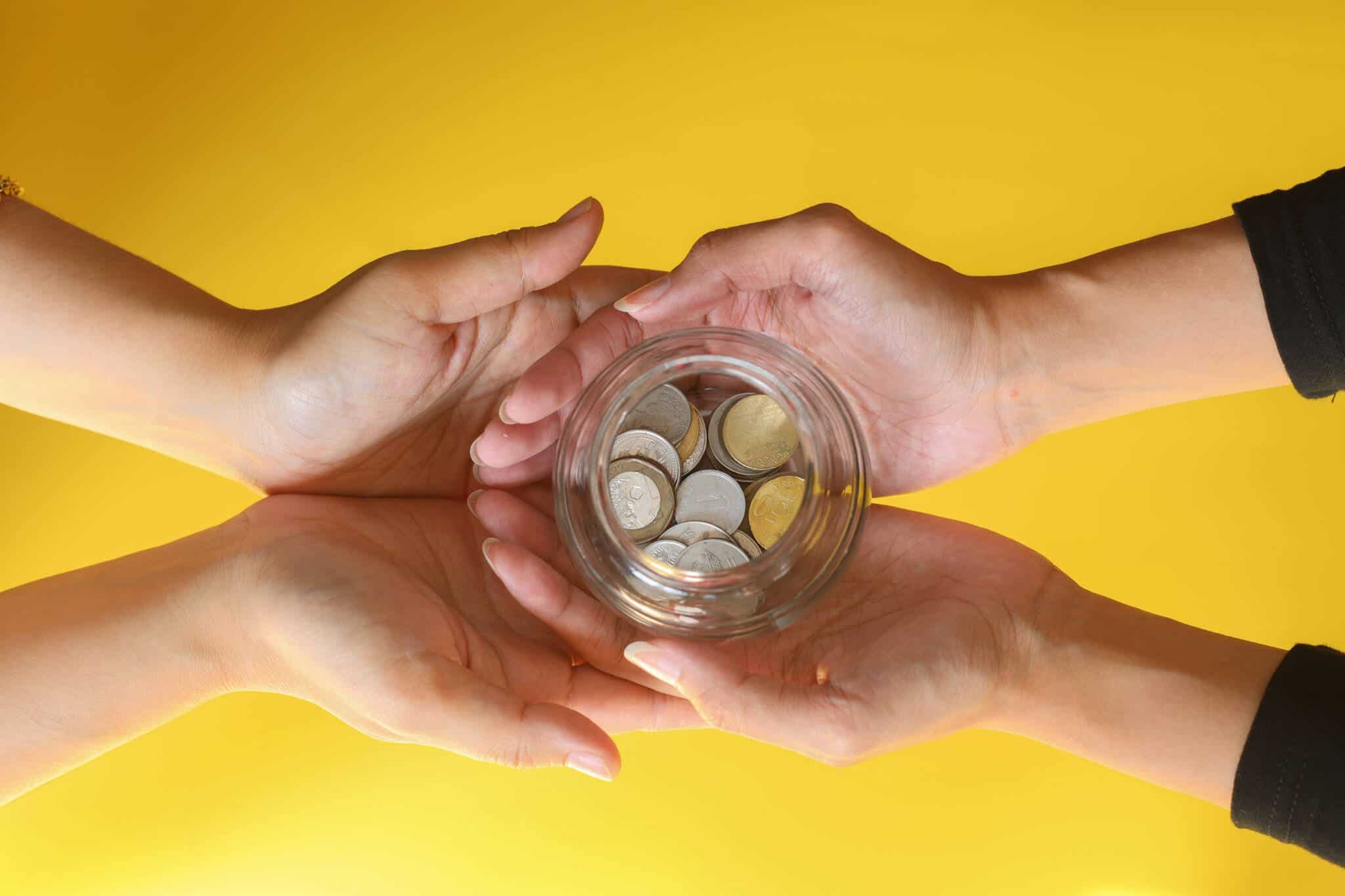 Mãos a oferecer frasco de vidro com moedas sobre fundo amarelo simbolizando utilizar IRS para ajudar
