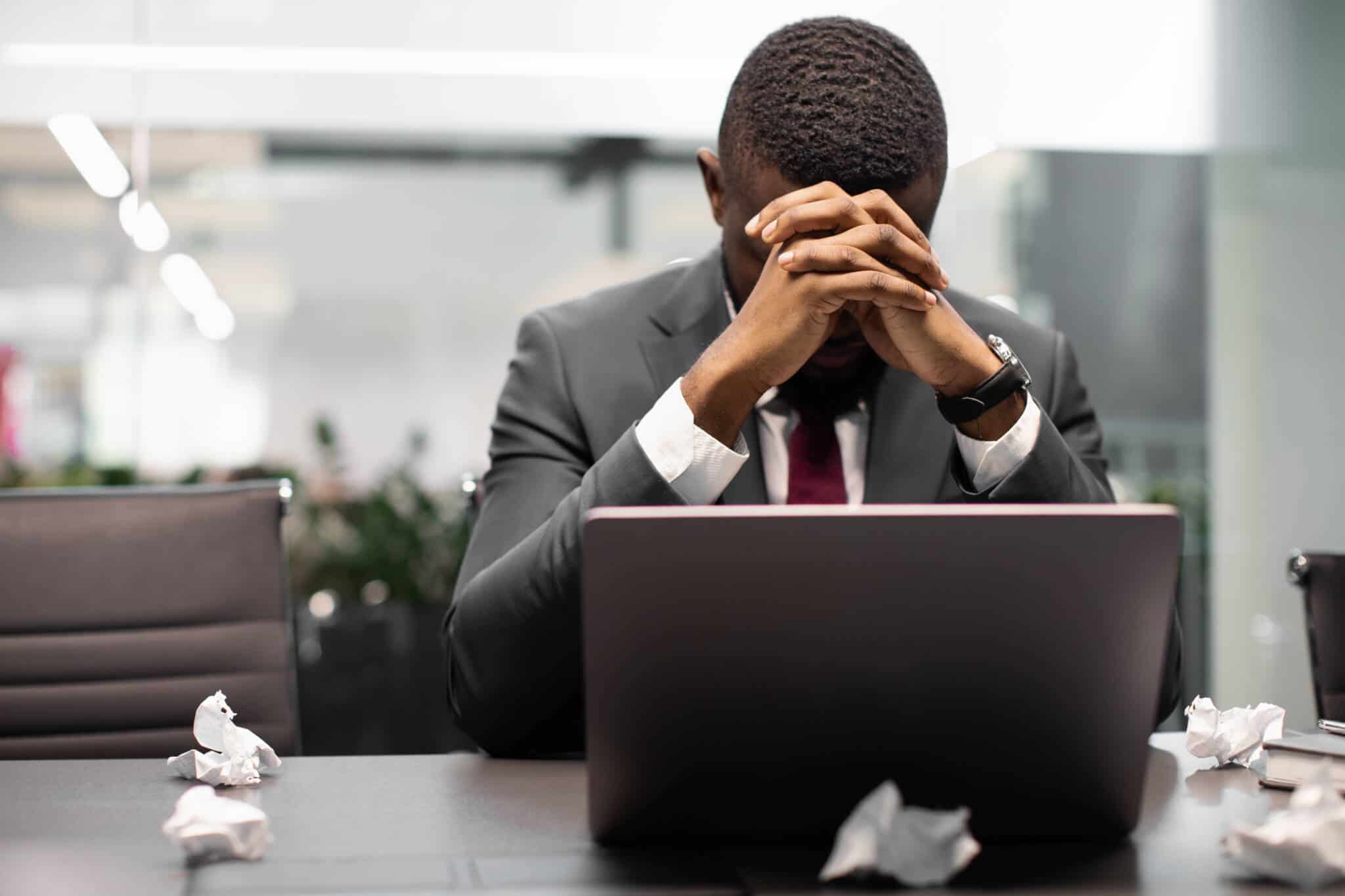 jovem homem, sentado à mesa do escritório, frente ao computador e rodeado de bolas de papel amachucado, está de mãos na cabeça mostrando-se em burnout