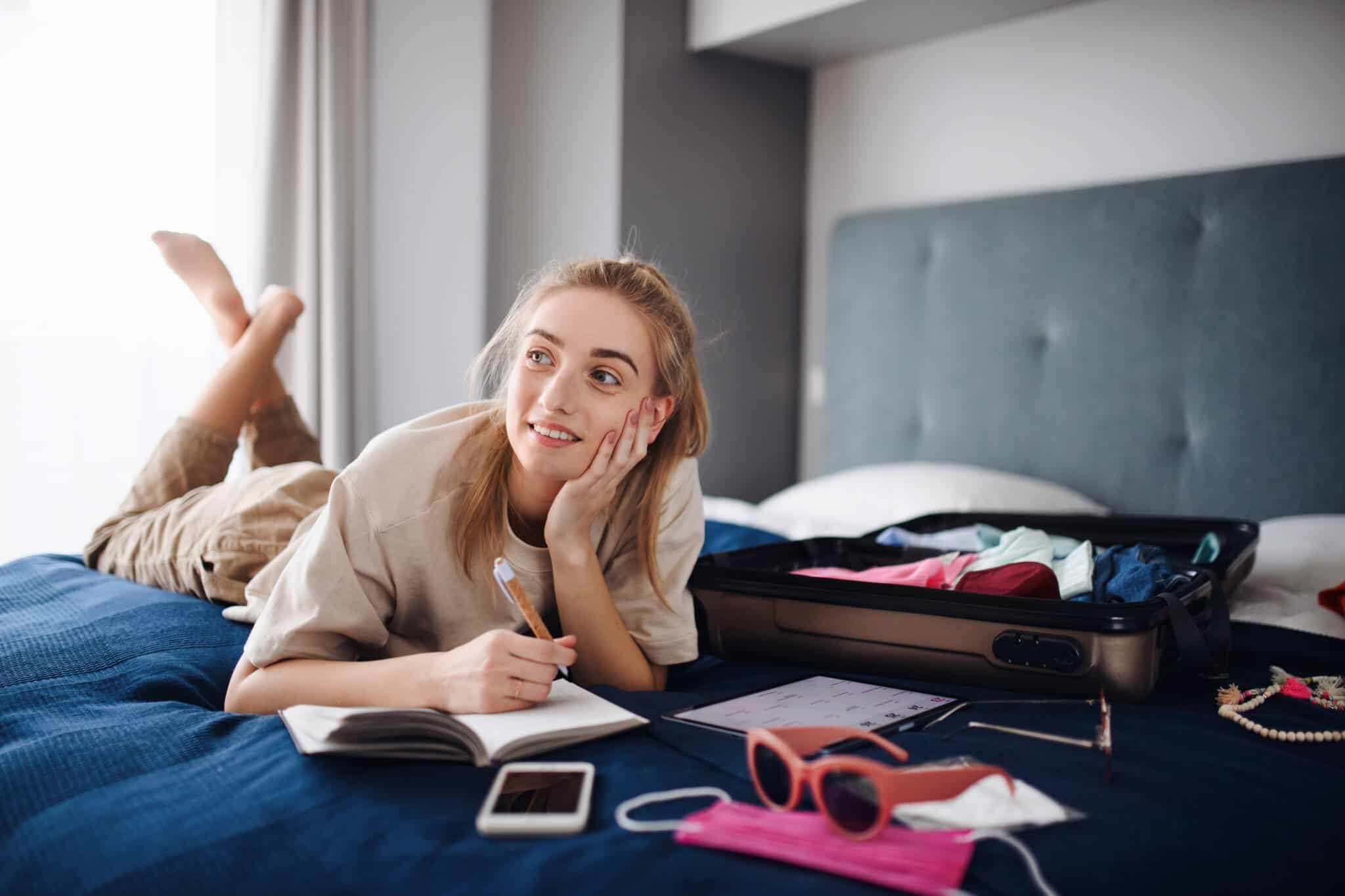 jovem mulher deitada na sua cama, onde tem a mala e vários objetos de viagem, enquanto planeia como poupar nas férias