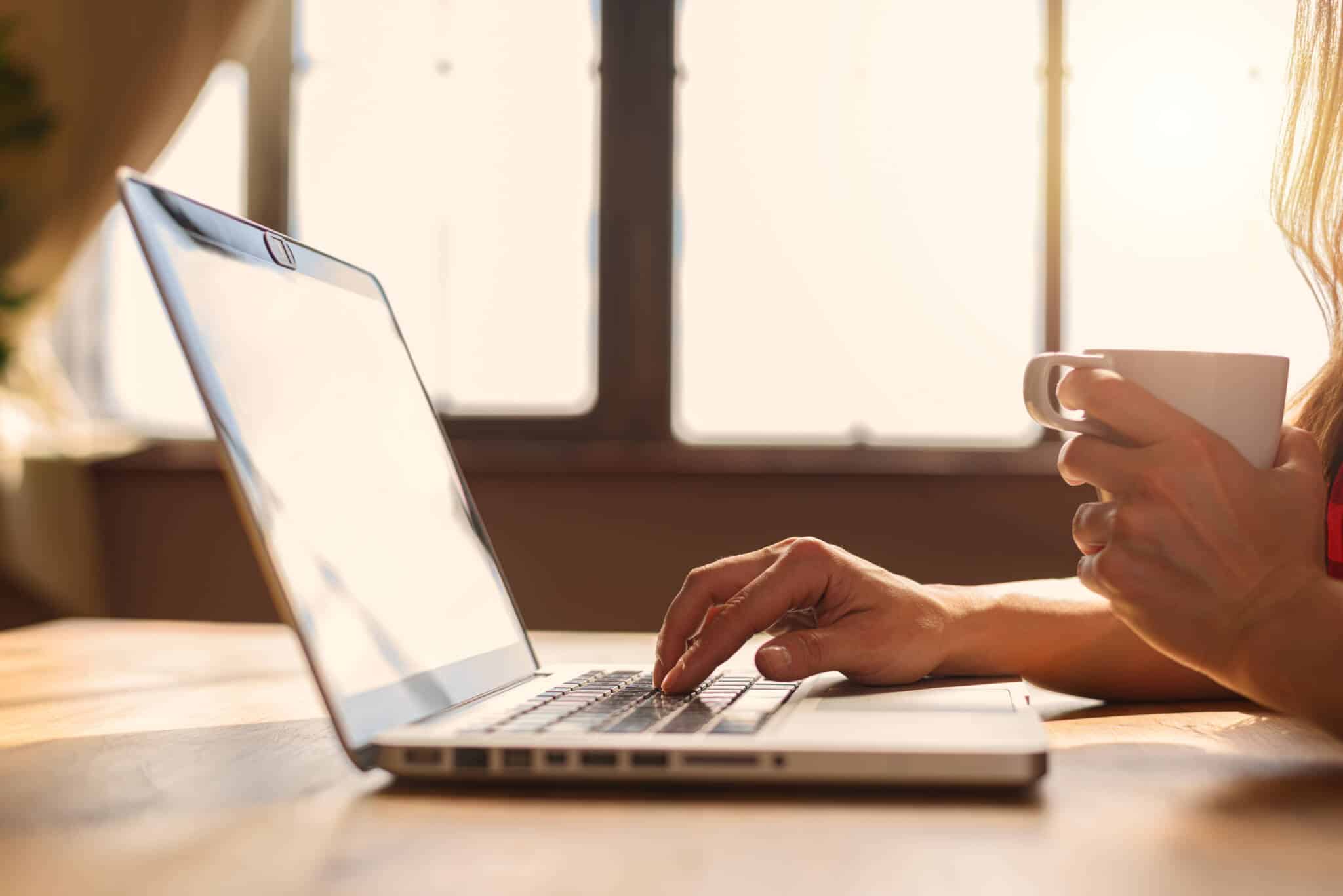 em contraluz, com um sol radiante, as mãos de uma jovem sentada a uma mesa junto a uma janela grande, usa o seu computador portátil usufruindo de teletrabalho