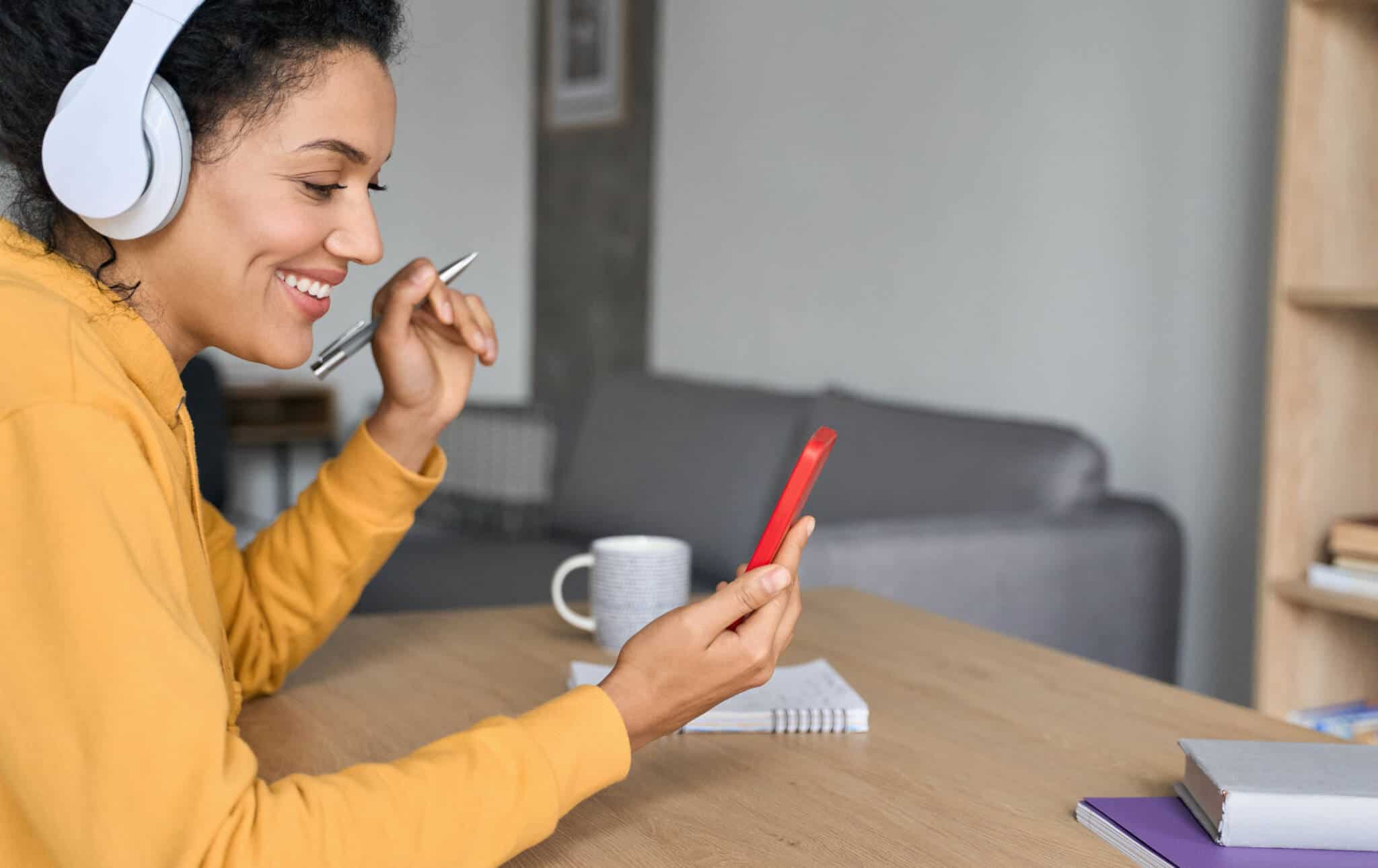 jovem mulher em casa, sentada à mesa da sala, ao telemóvel usa aplicação para aprender inglês, tirando notas para um bloco de papel