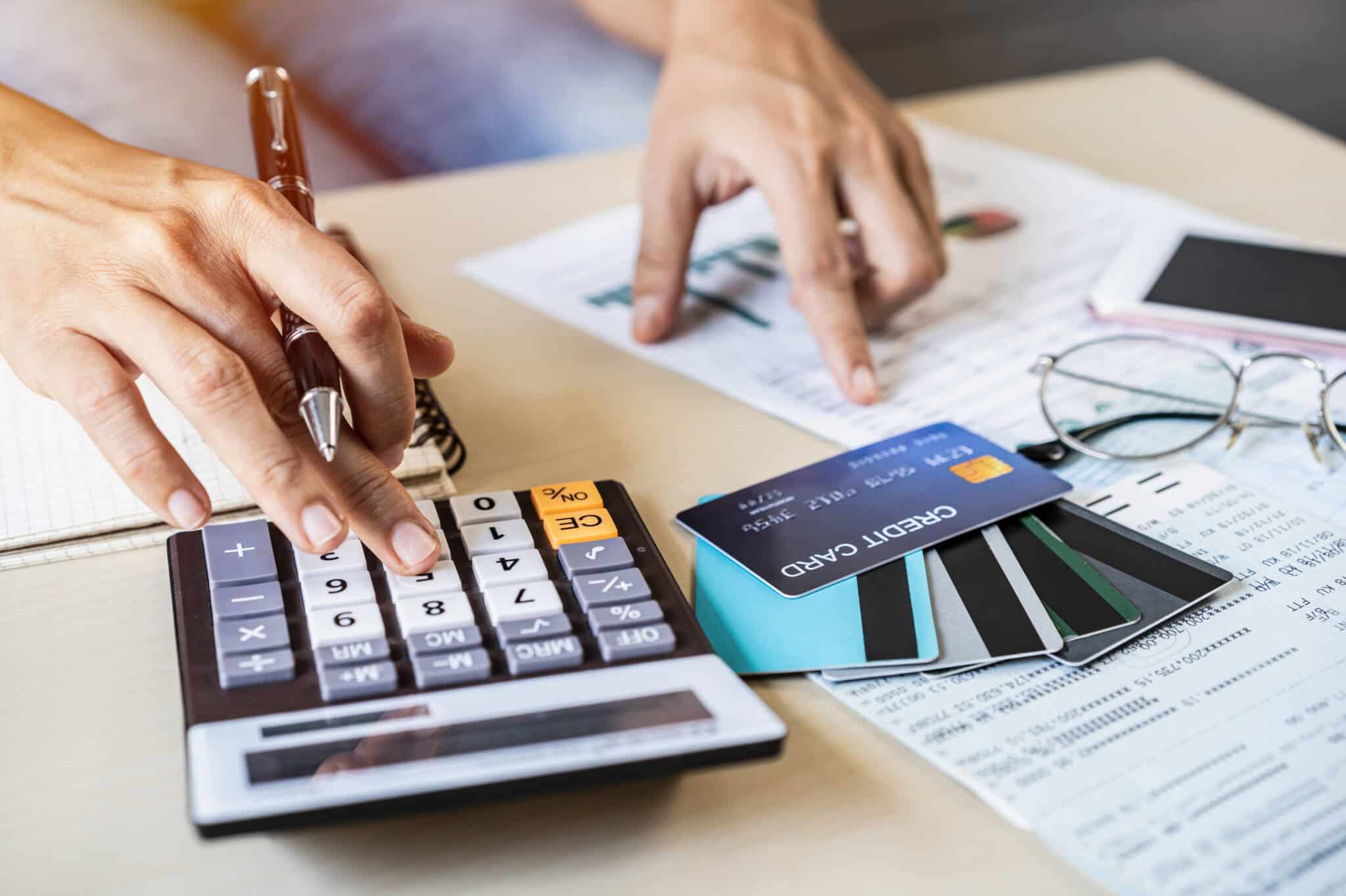 mão de jovem mulher a fazer contas na calculadora e analisar documentos espalhados na mesa onde estão também um conjunto de cartões bancários, produtos de contas bancárias