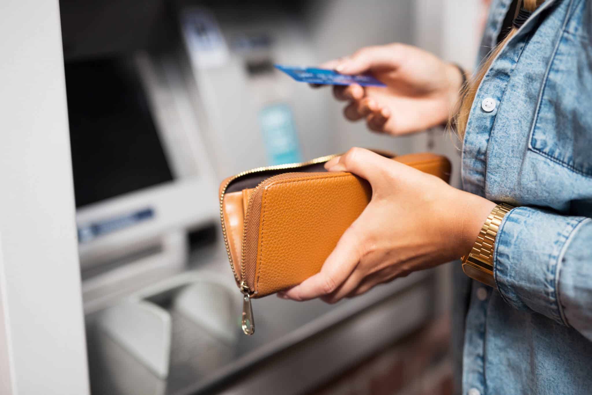 jovem mulher junto a um ATM segura cartão retirado da carteira aberta para introduzir e fazer uma transferência pelo multibanco